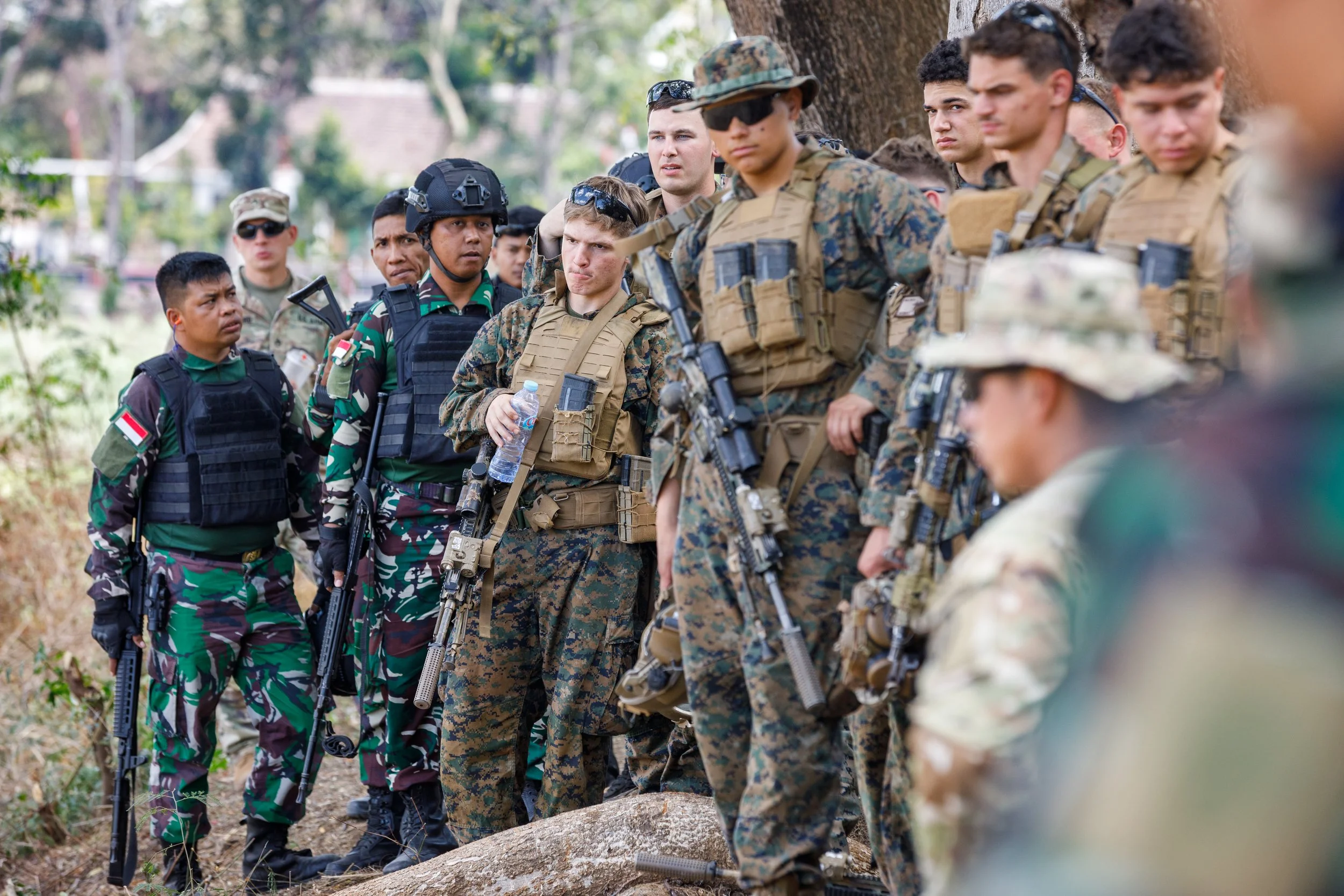 Group of soldiers in military uniform and gear, standing outdoors near a tree, listening intently.