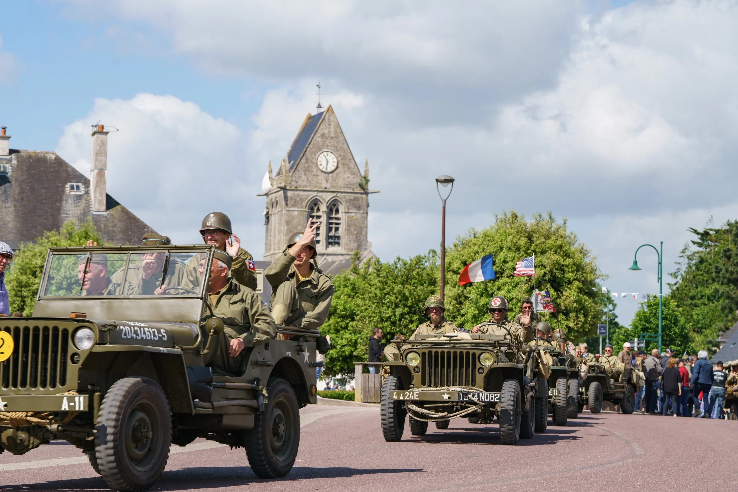 Military vehicles and soldiers participating in a parade, with people watching, historic buildings, and flags in the background.