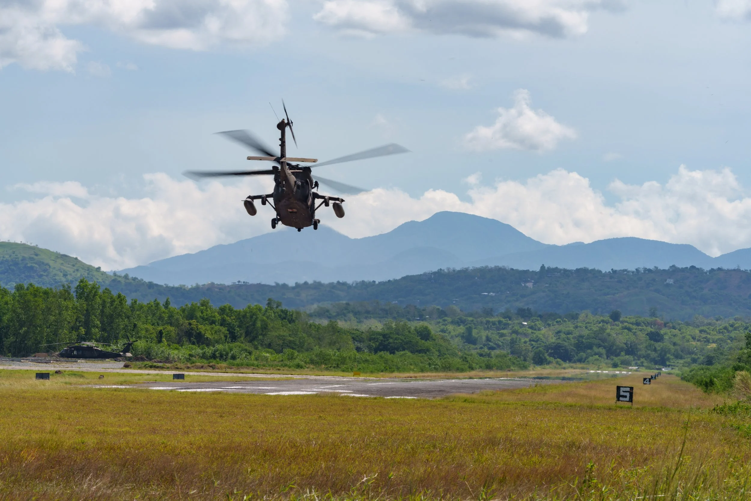 A military helicopter flying above a grassy field with another helicopter on the ground and mountains in the background.