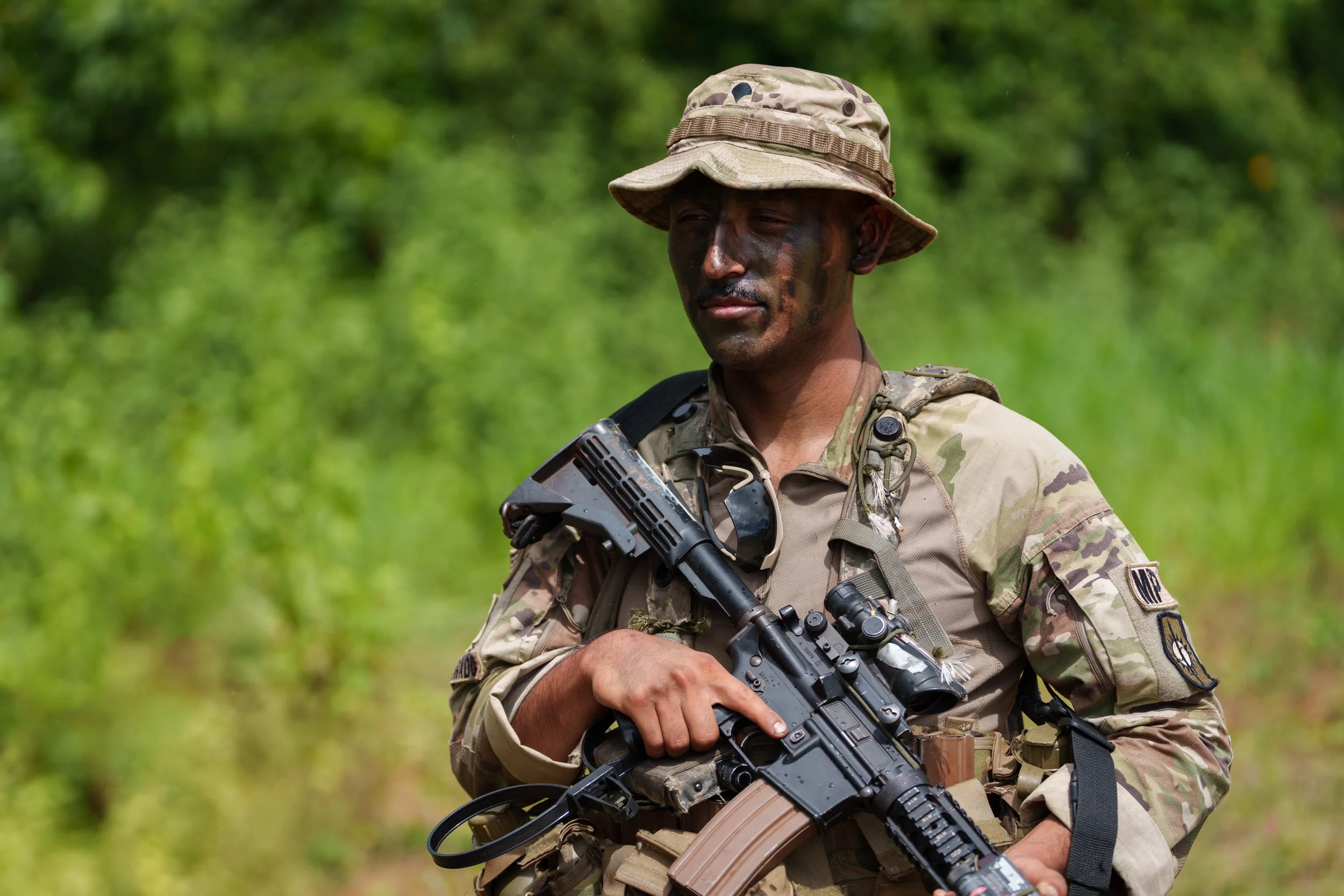 A soldier in camouflage uniform holding a rifle outdoors with green foliage in the background.