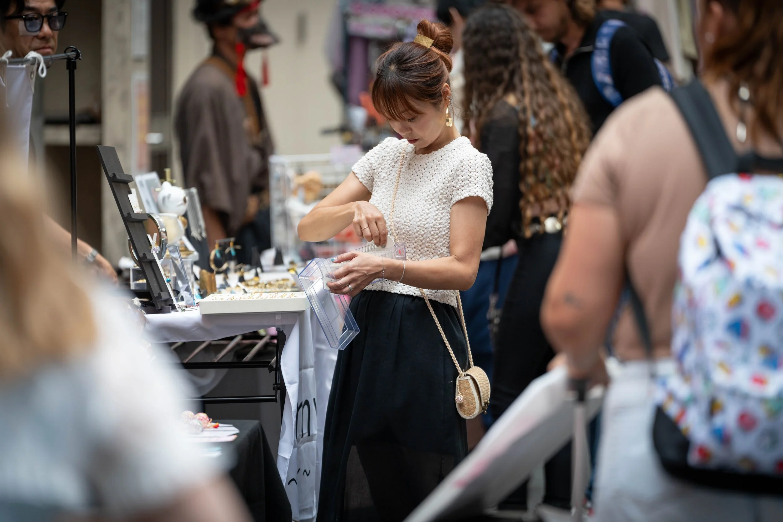 A woman shopping at a jewelry booth during a market or fair, looking at jewelry displayed on the table.