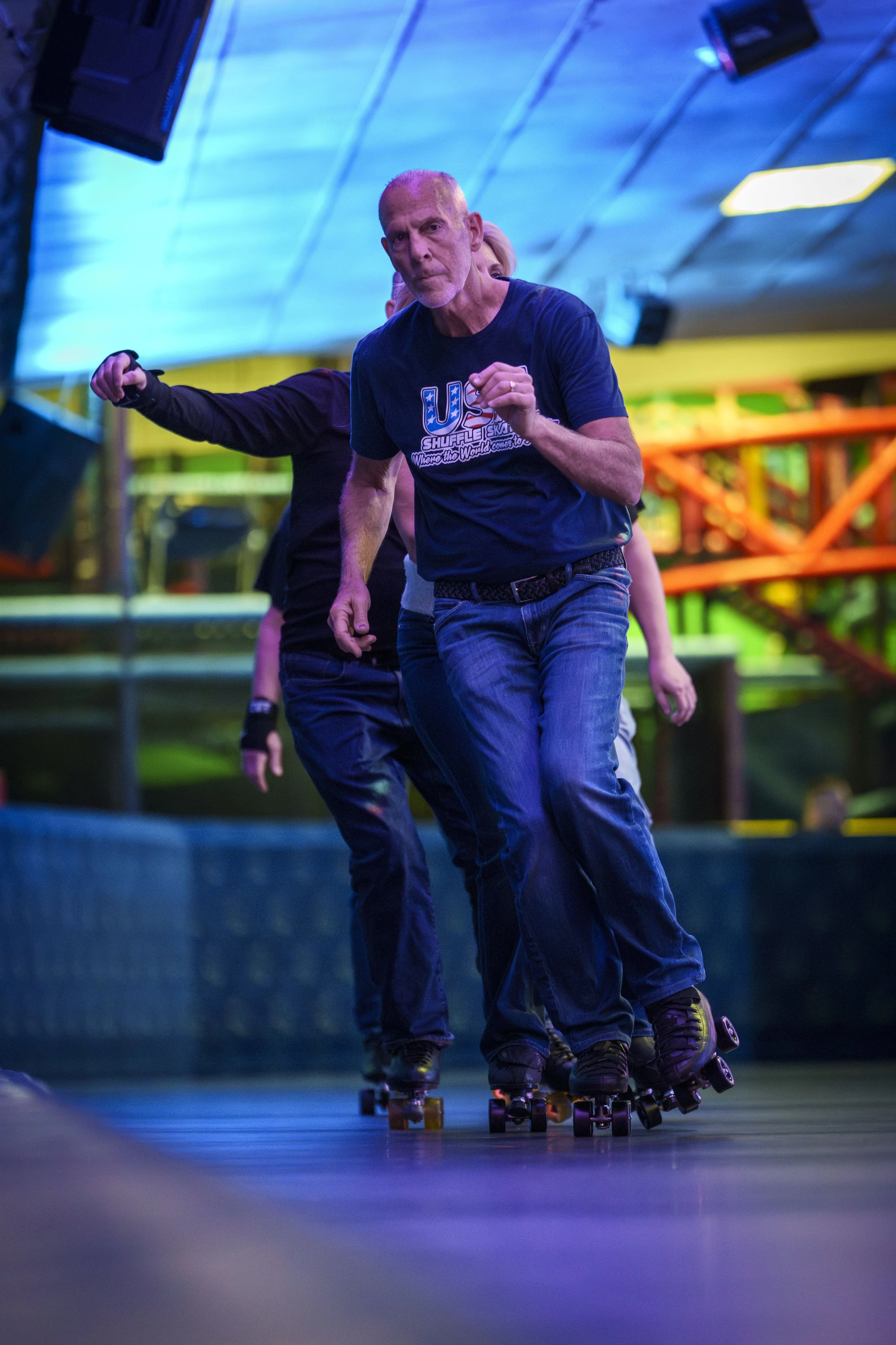 Older man roller skating indoors with others, colorful lighting, and vibrant background.
