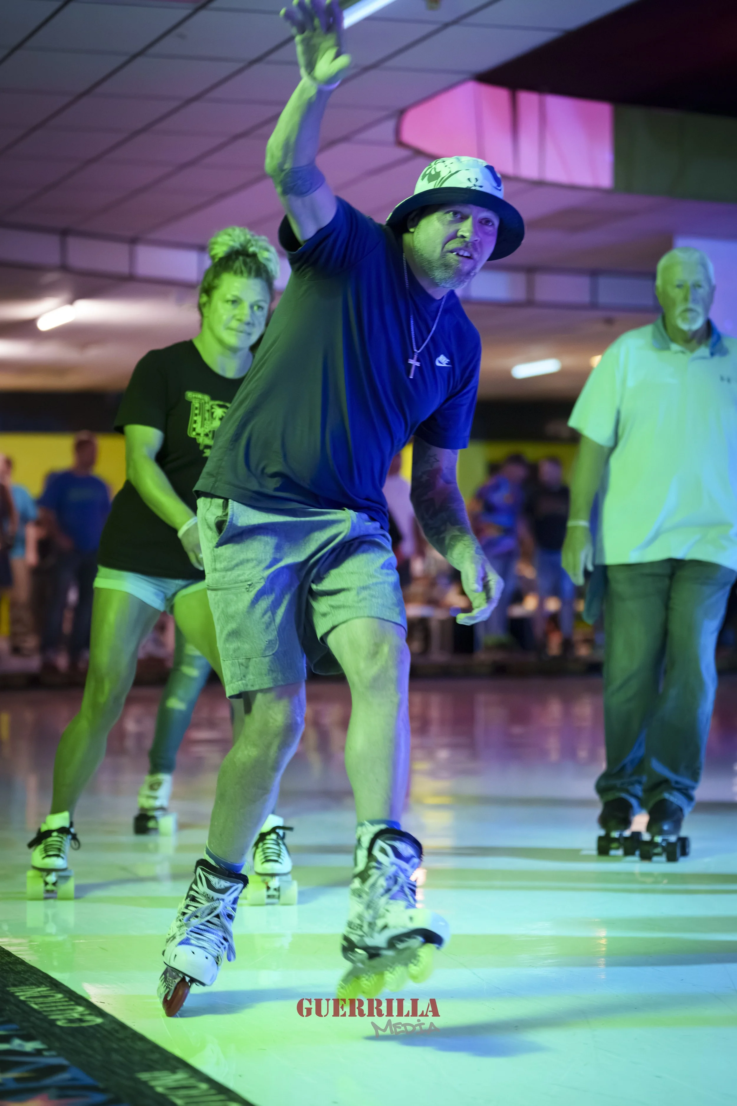 People roller skating indoors, with one man in a hat and cross necklace waving at the camera, under colorful lighting.