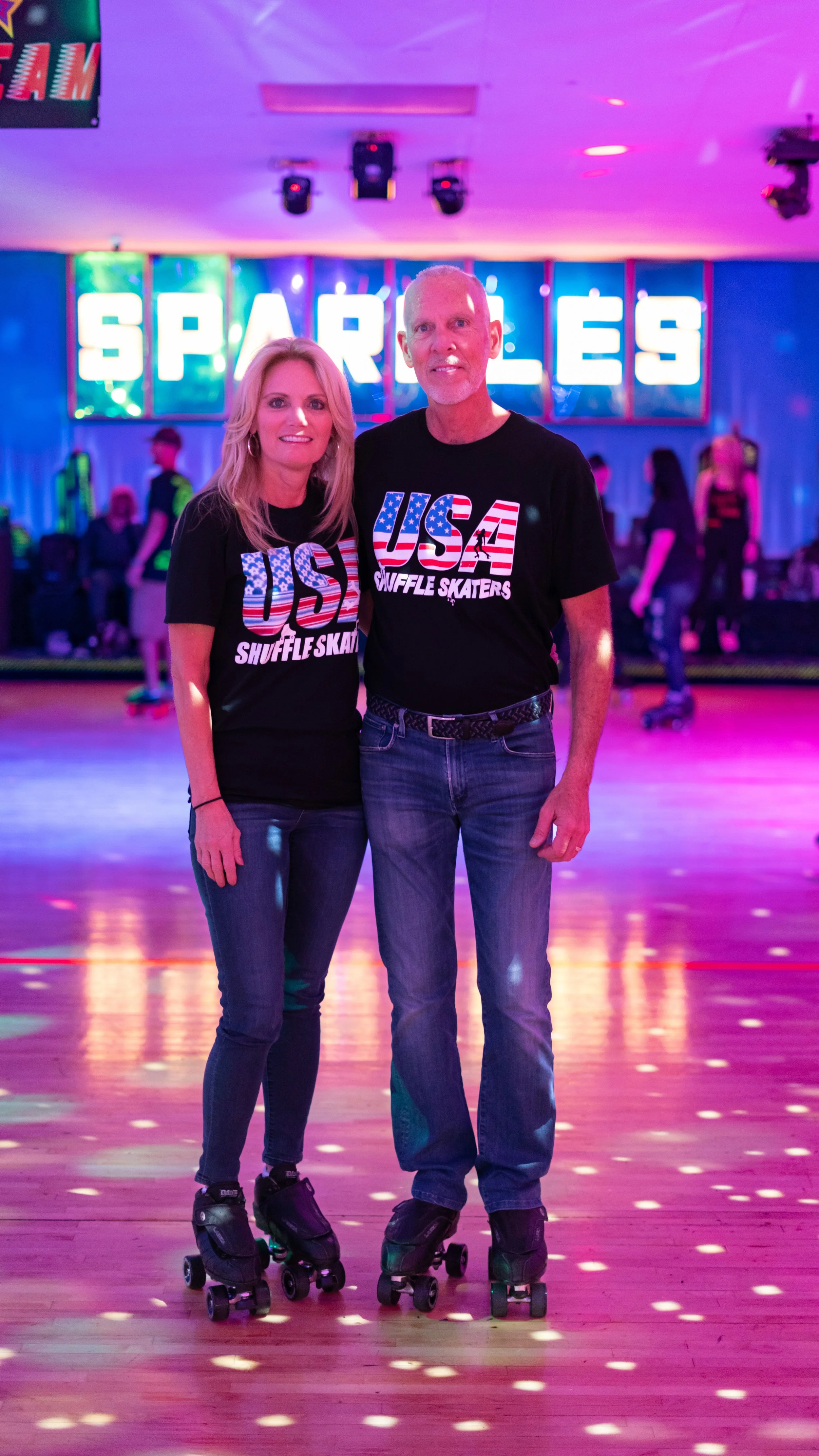 A man and woman wearing matching USA shuffle skaters T-shirts, roller skating indoors with a colorful, neon-lit background that says 'SPARKLES,' and other people skating in the background.