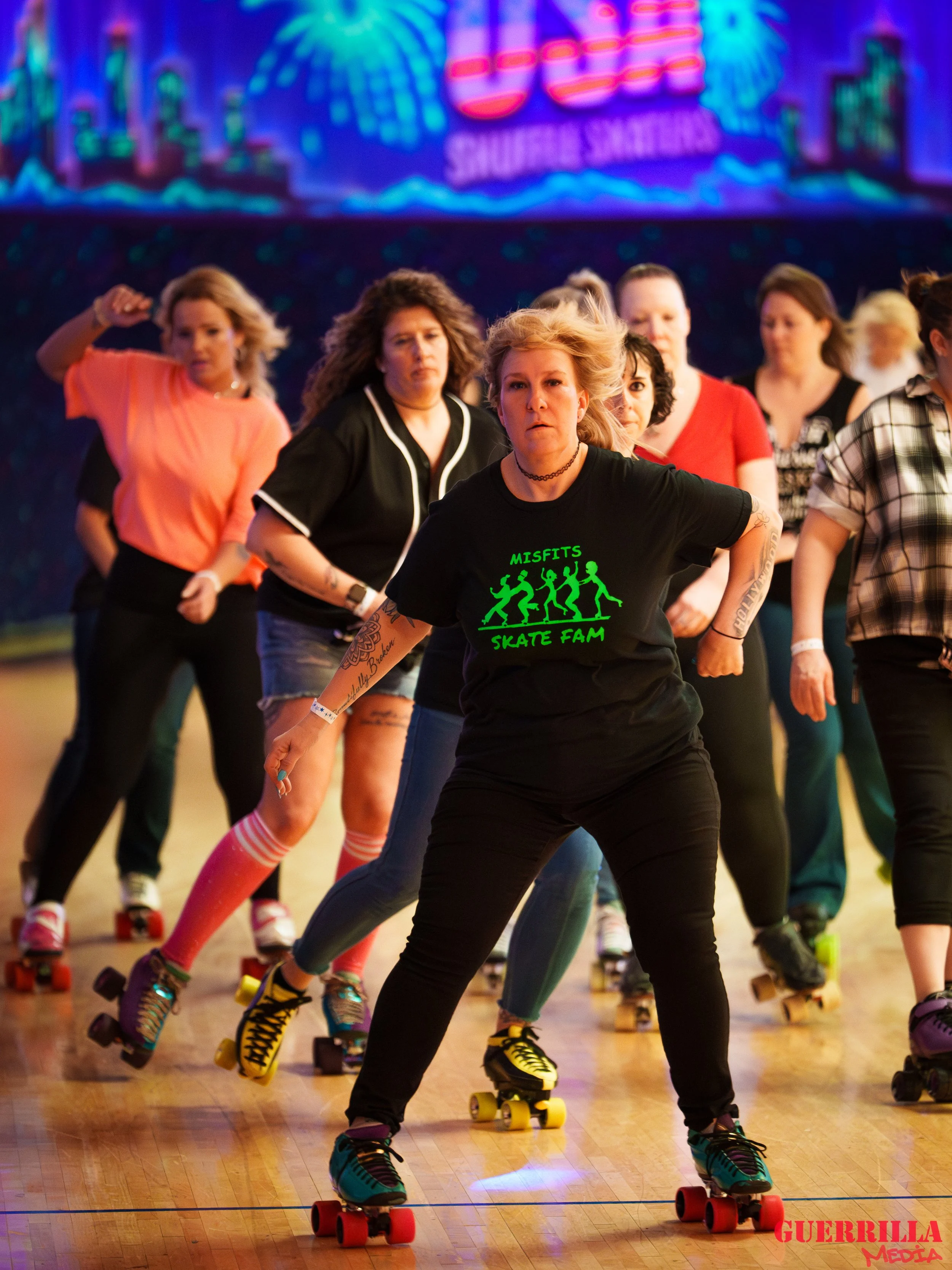 Group of women roller skating in a large indoor space, with a neon sign in the background, some wearing casual clothes and skate shoes, others wearing themed T-shirts.