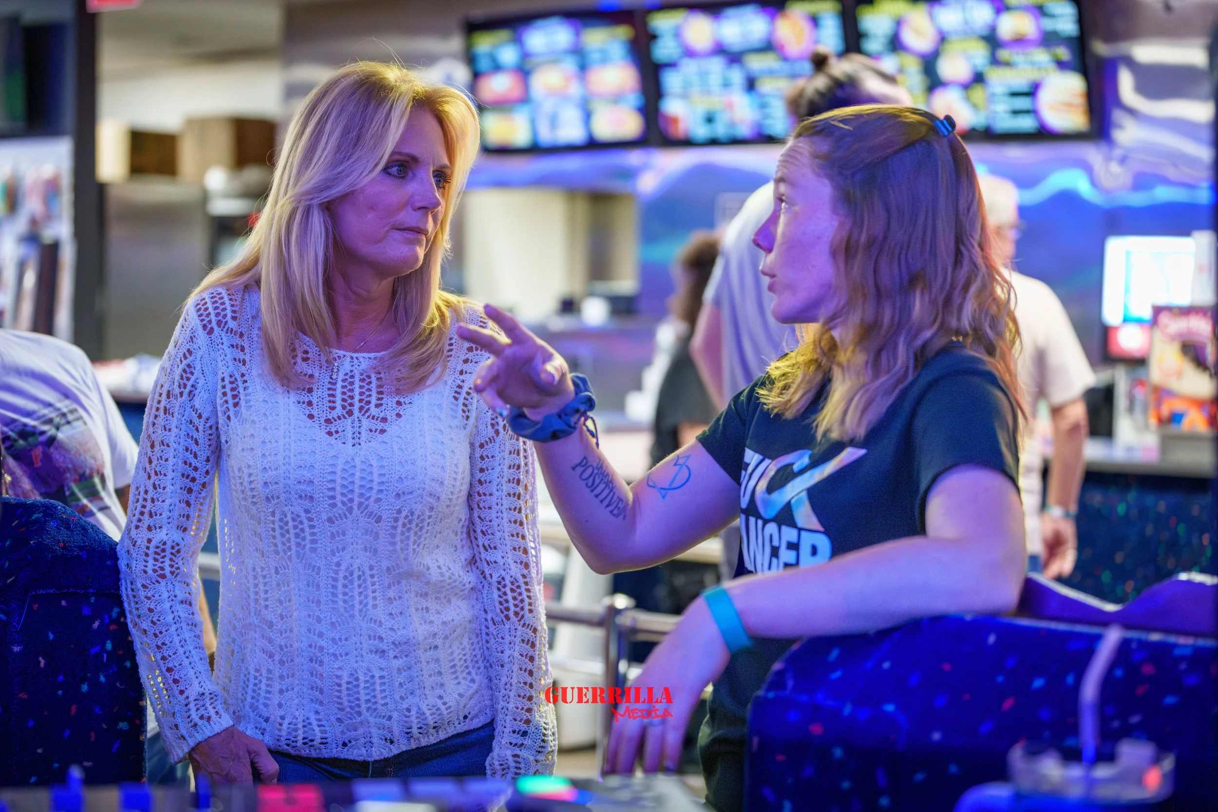 Two women are having a serious conversation in a colorful arcade or gaming lounge. The woman on the left has blonde hair and is wearing a white lace sweater, looking concerned. The woman on the right has light brown hair with roots visible, wearing a