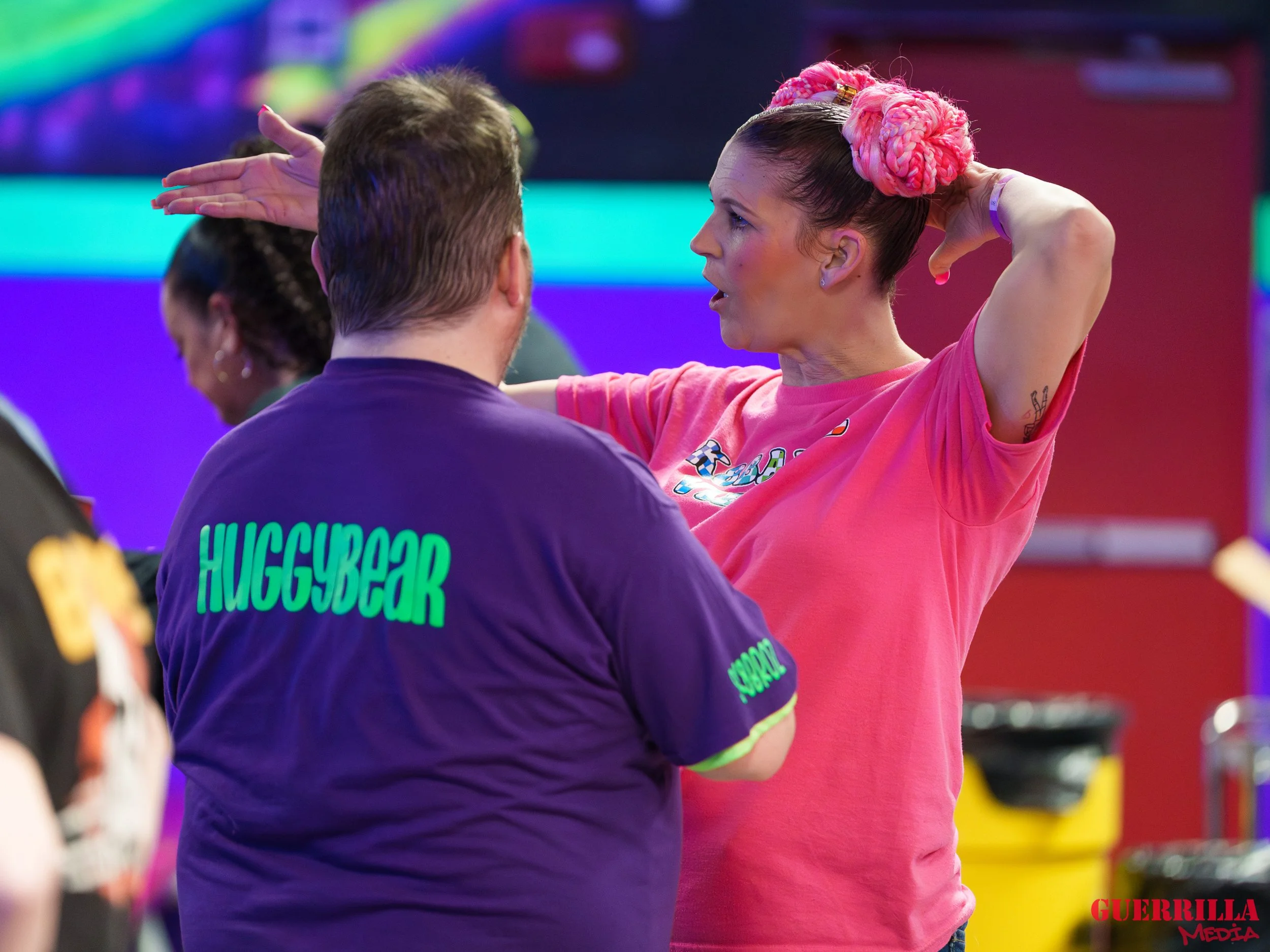 A woman with pink braided hair and a pink t-shirt, gesturing with her hands, appears to be speaking to a man with a navy shirt that has “HUGGYBEAR” written on the back. They are in an indoor setting with colorful lights.