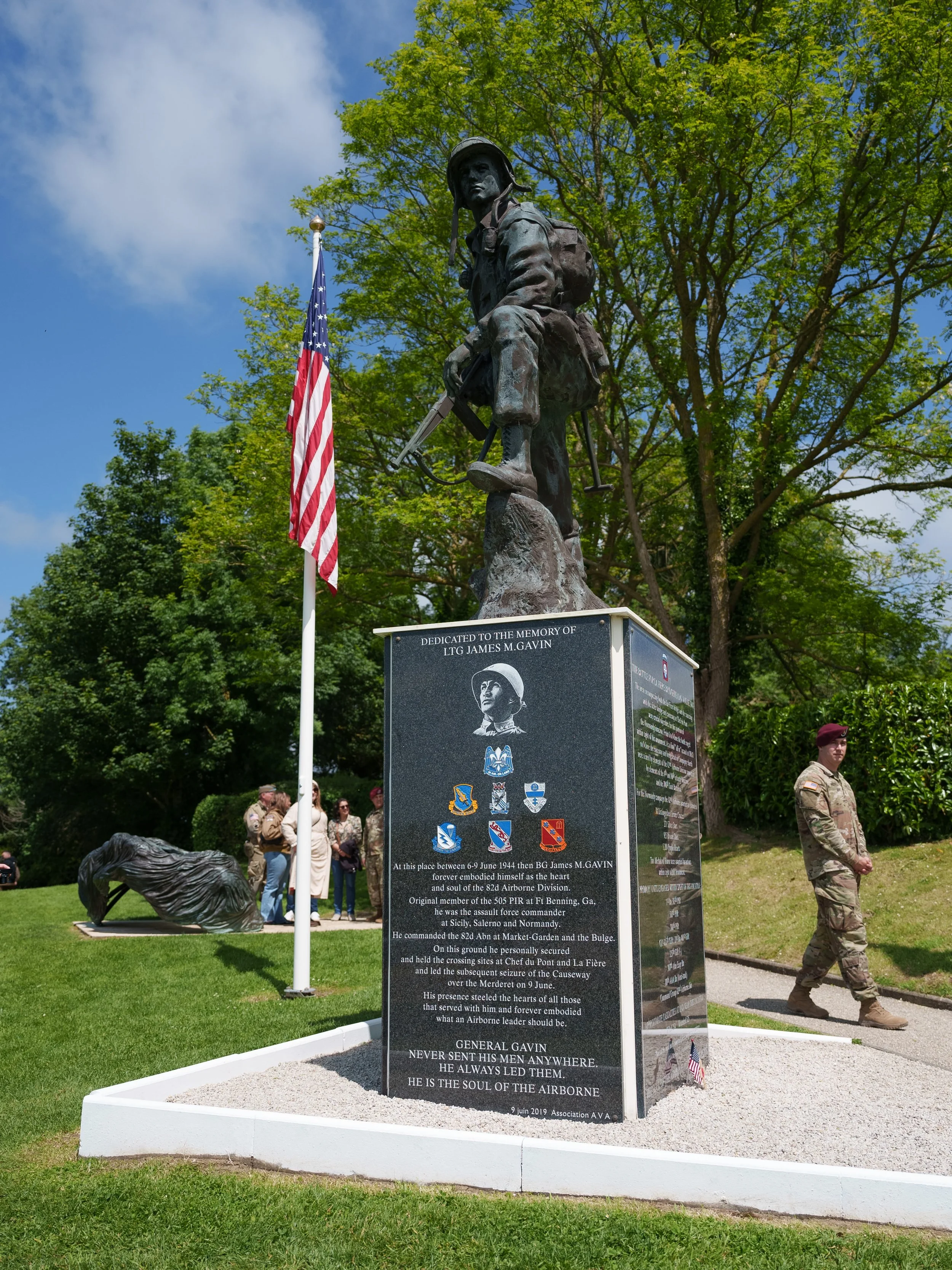 A memorial with a bronze statue of a soldier holding a rifle, standing on a pedestal with engravings and military insignias. An American flag is nearby, and people in military uniform and civilians are gathered around, with trees and a blue sky in th