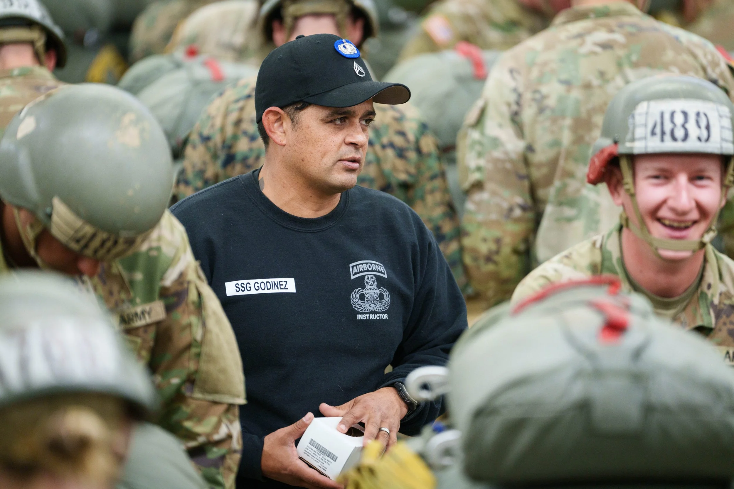 A military instructor labeled "SSG Godinez" teaches soldiers, all in camouflage uniforms and helmets, in a classroom or briefing setting.