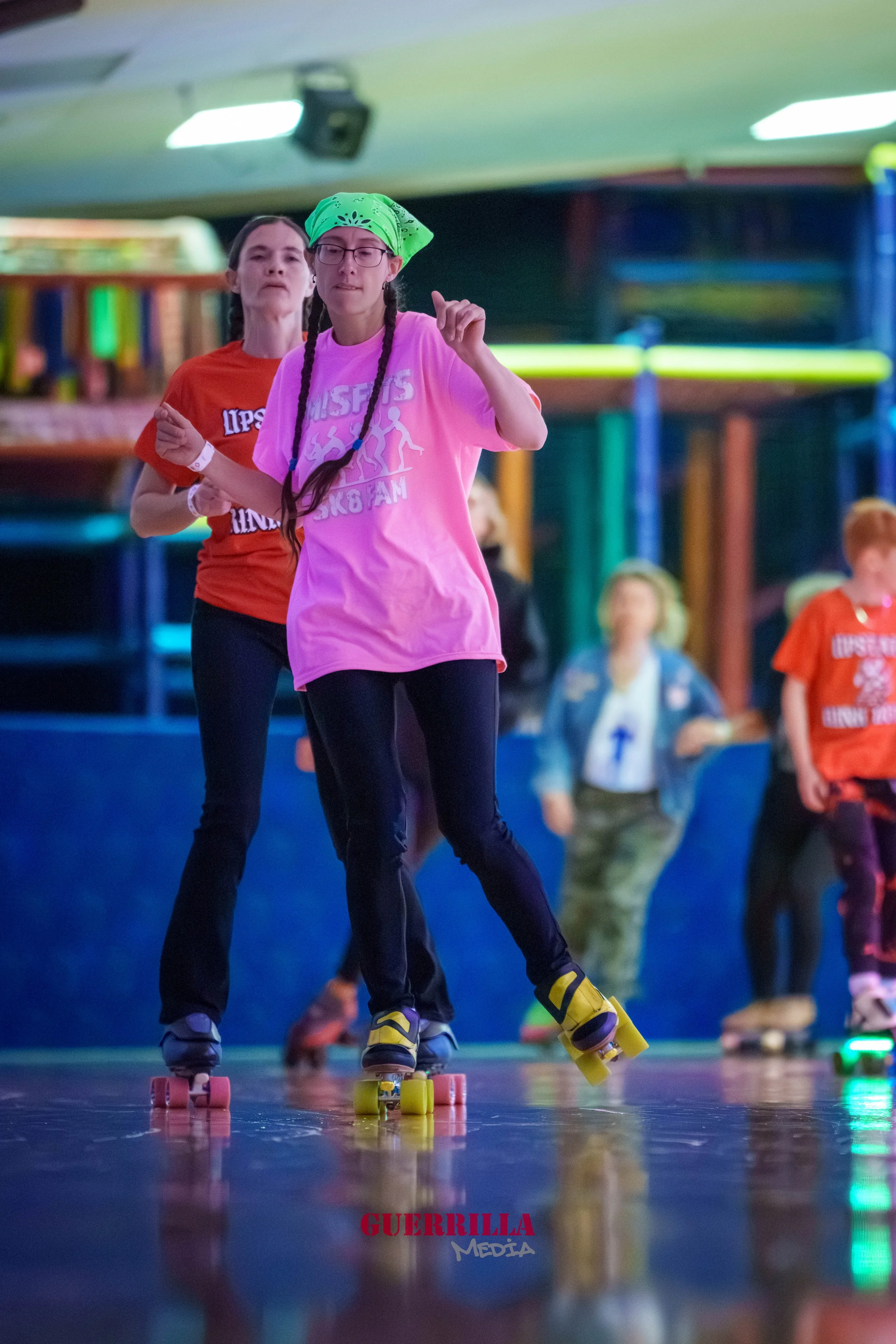 Three girls roller skating indoors with colorful background. One girl in pink in the front, another in orange behind, and a third in a blue jacket visible in the background.