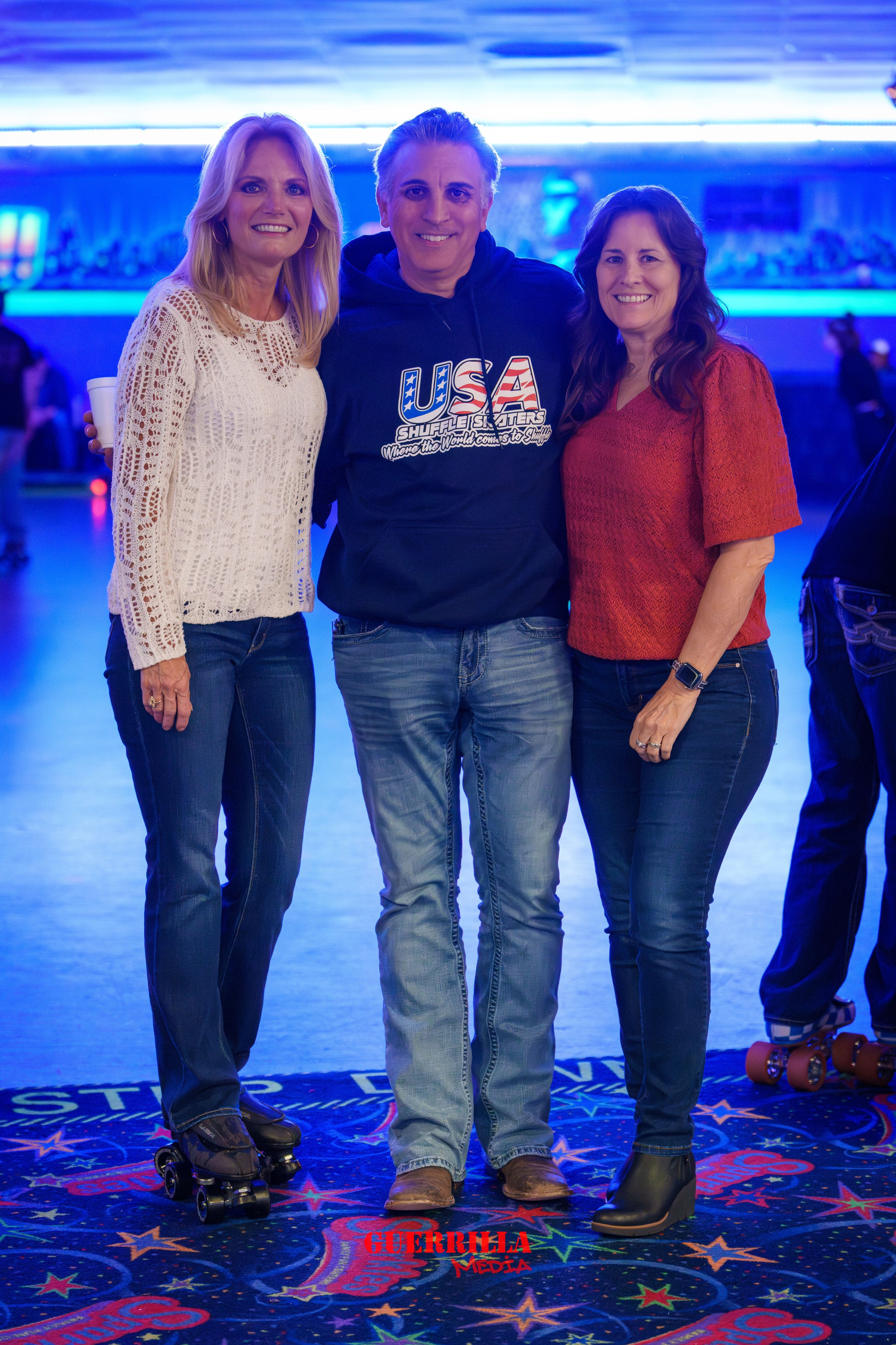 Three people standing together at a roller skating rink with a blue-lit background, the woman on the left is on roller skates, and the woman on the right is wearing a smartwatch. All three are smiling.