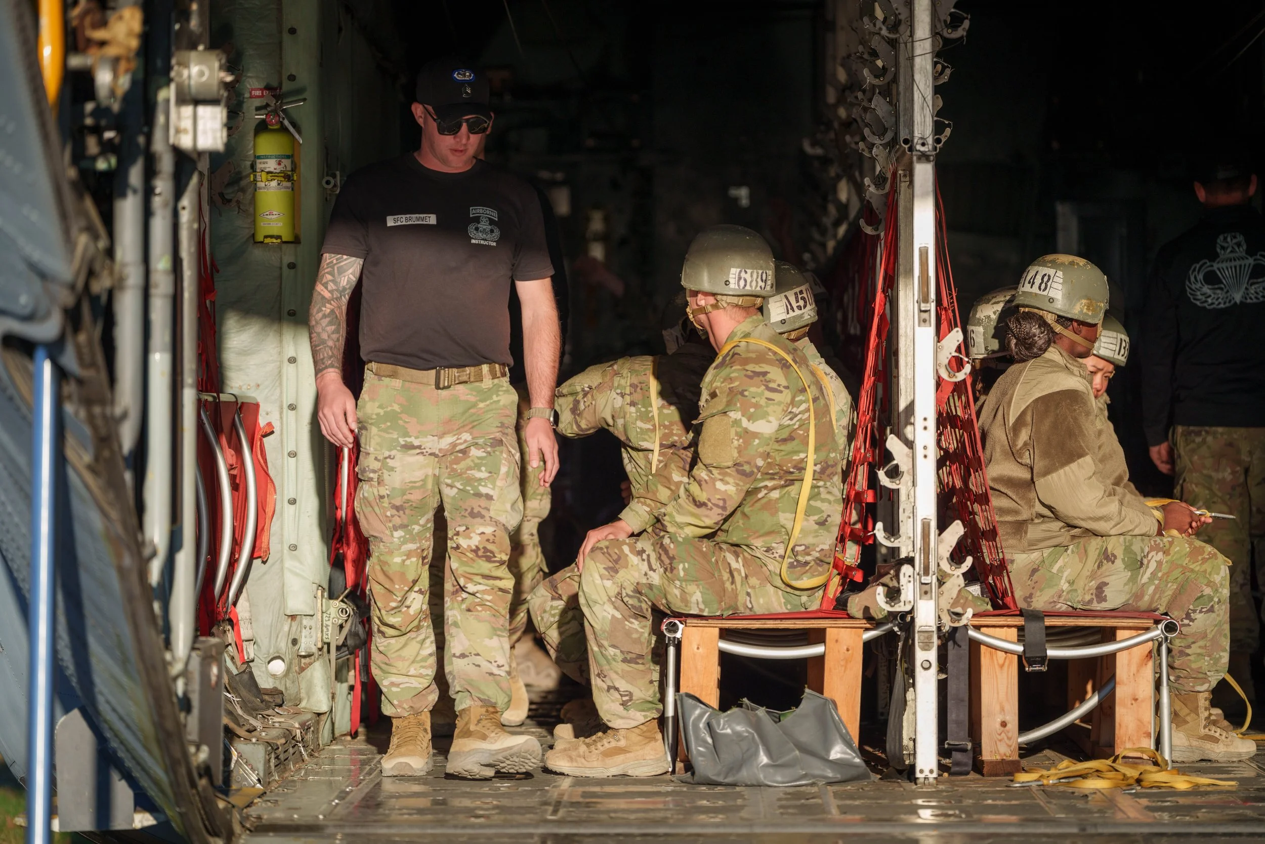 Military personnel in uniform inside a cargo aircraft, some seated, some standing, with equipment and safety gear.