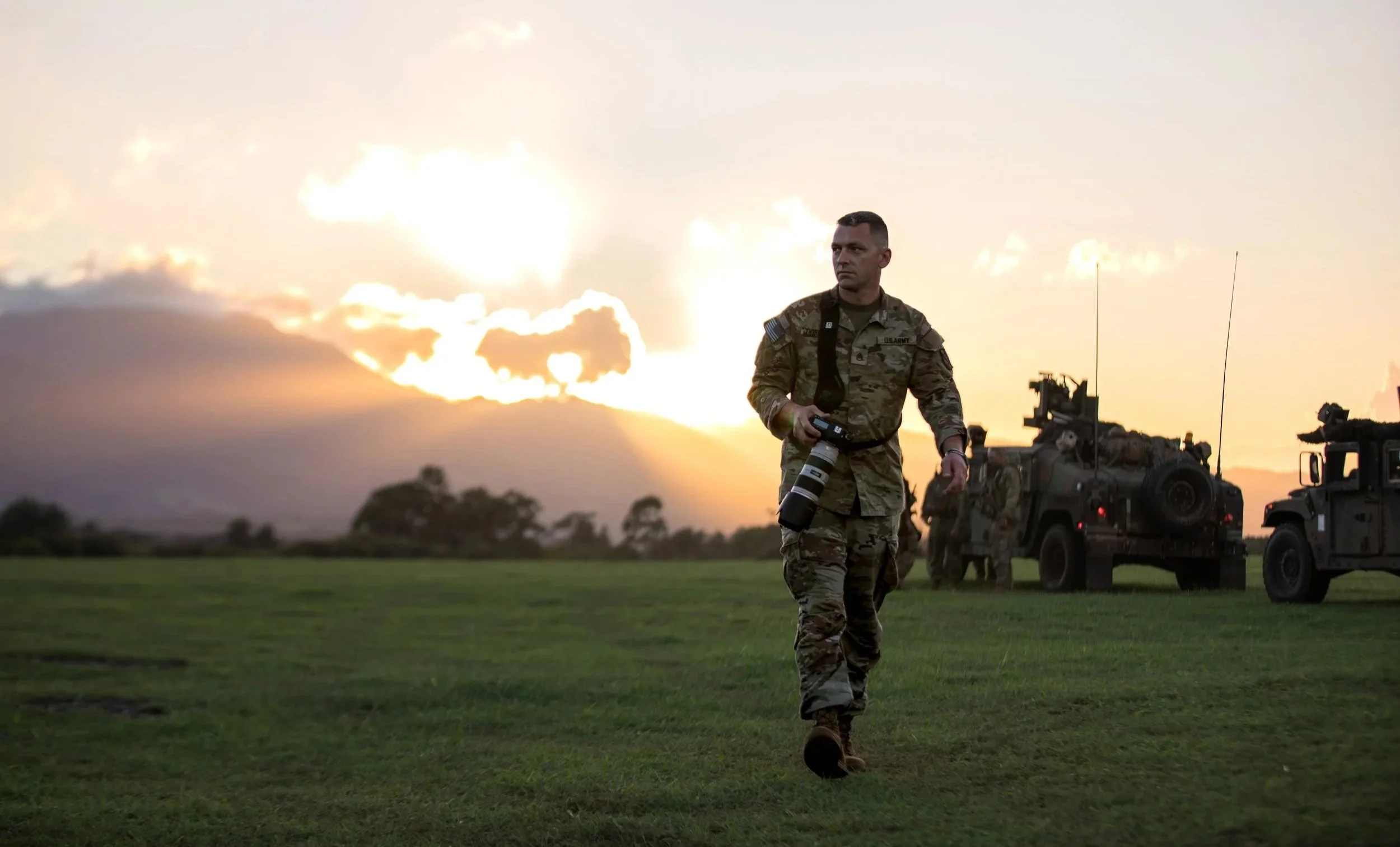 A soldier in camouflage military uniform walking across a grassy field at sunset with military vehicles in the background.