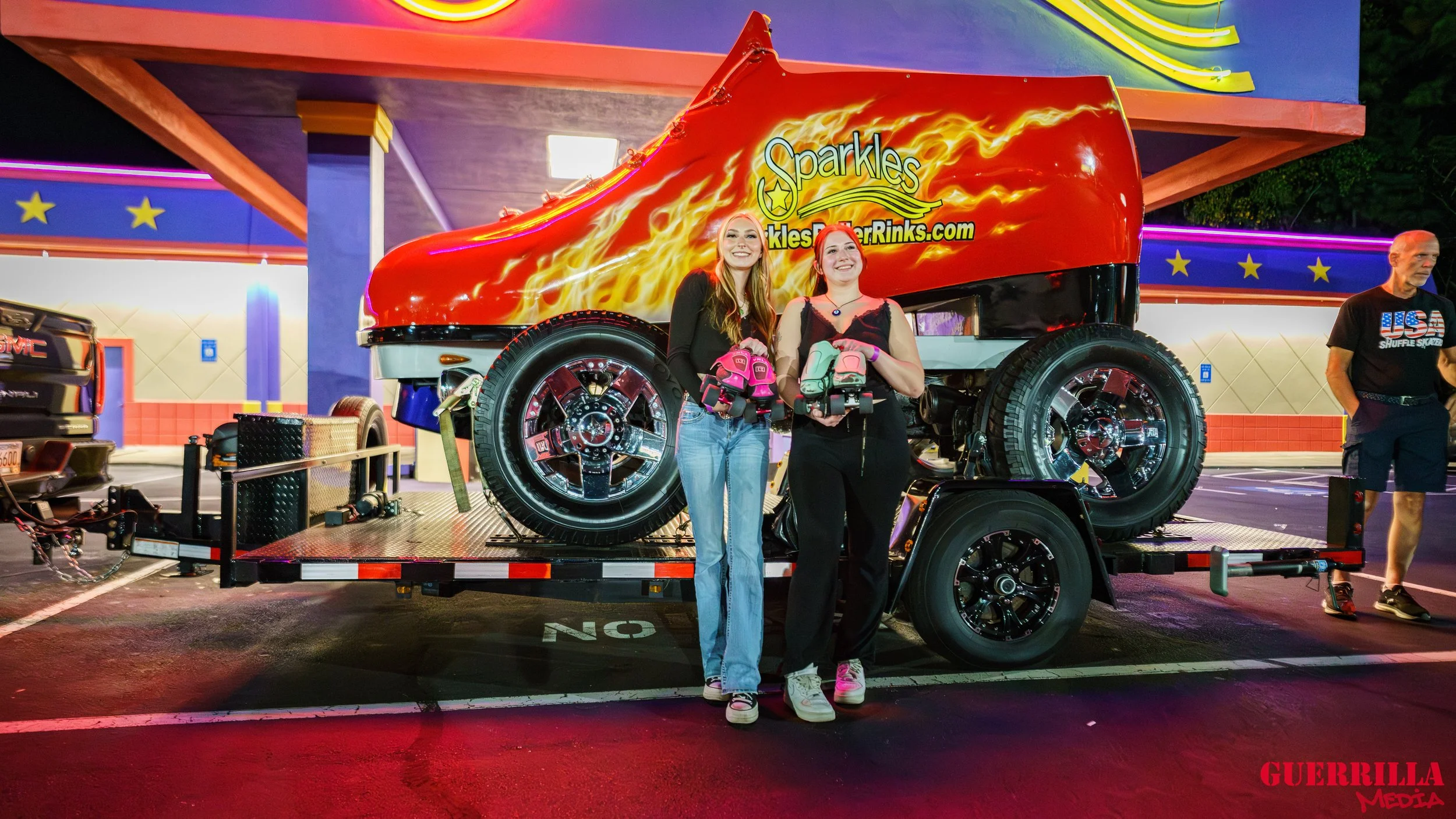 Two women standing next to a monster truck decorated with flames and the word 'Sparkles' with sparkles and a website address, holding roller skates, at a carnival or fairground at night.