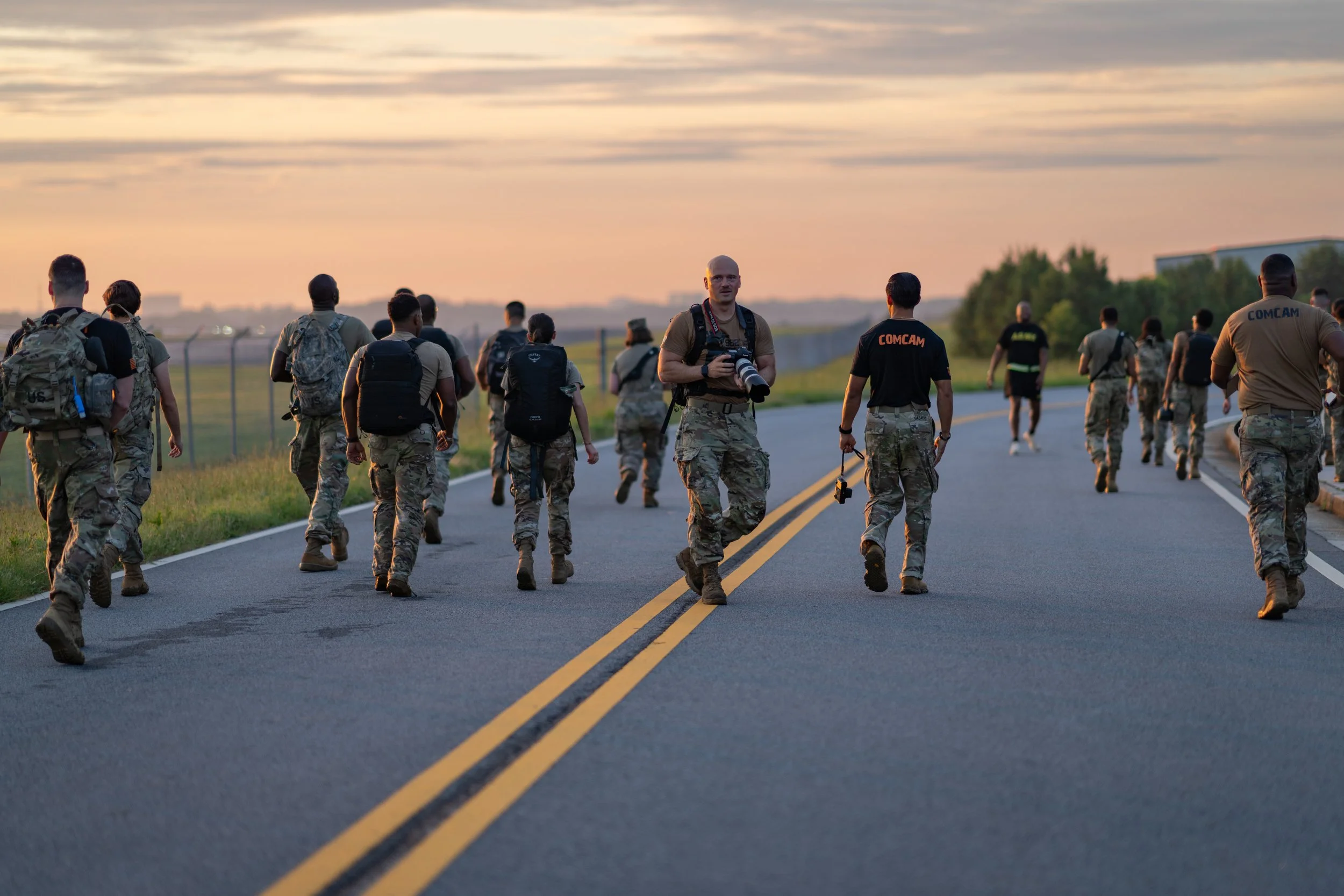 Group of military personnel walking along a deserted road at sunset, some carrying backpacks and cameras.