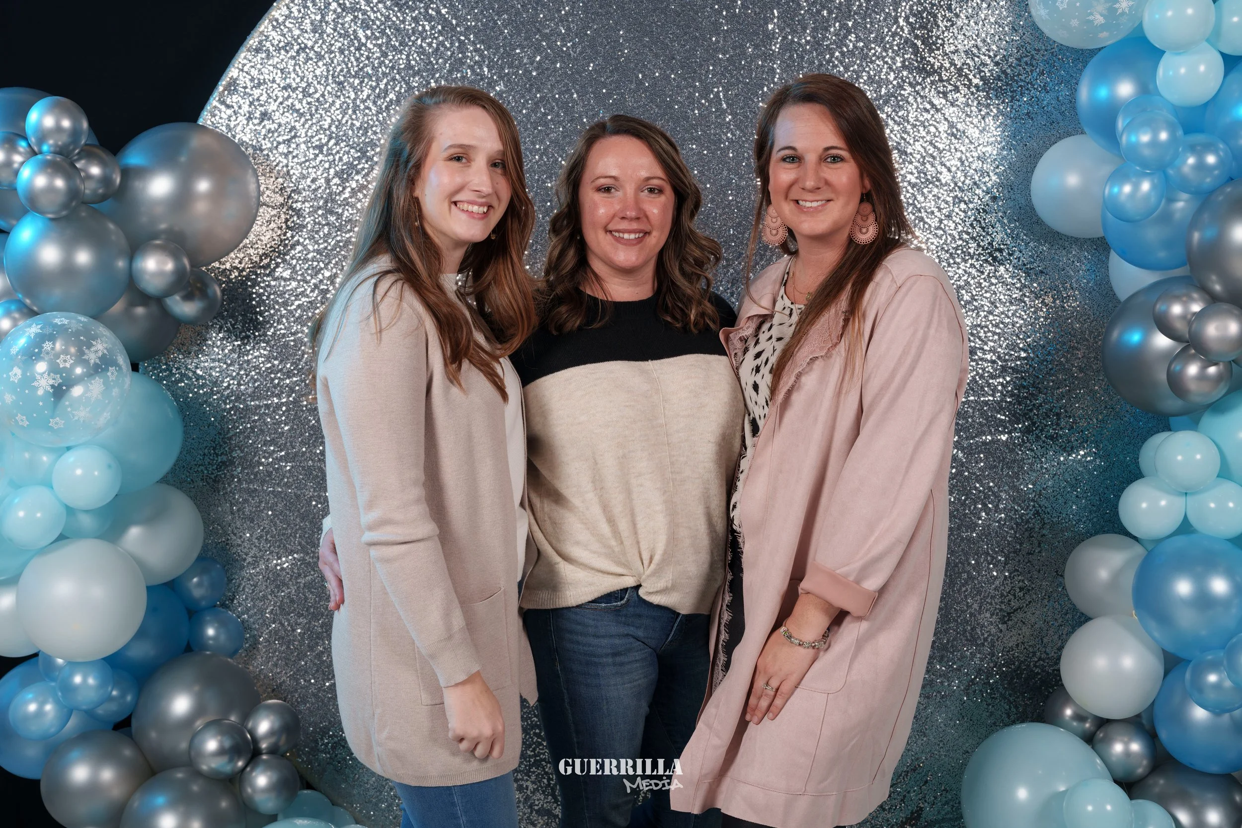 Three women posing in front of a silver glitter backdrop with balloon arch made of blue, silver, and white balloons.