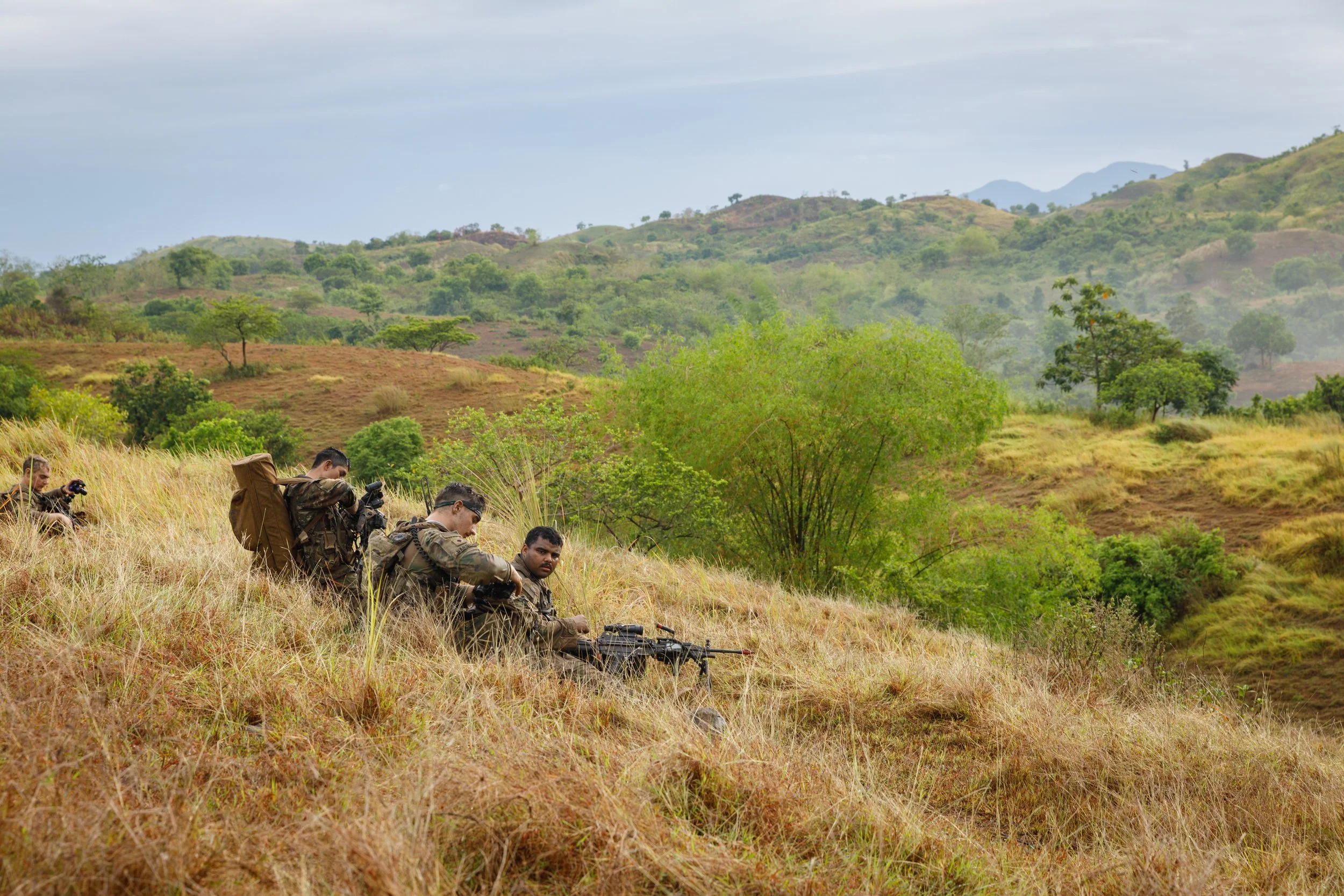 A group of soldiers in camouflage uniforms resting in tall grass on a hillside, with green trees and rolling hills in the background.