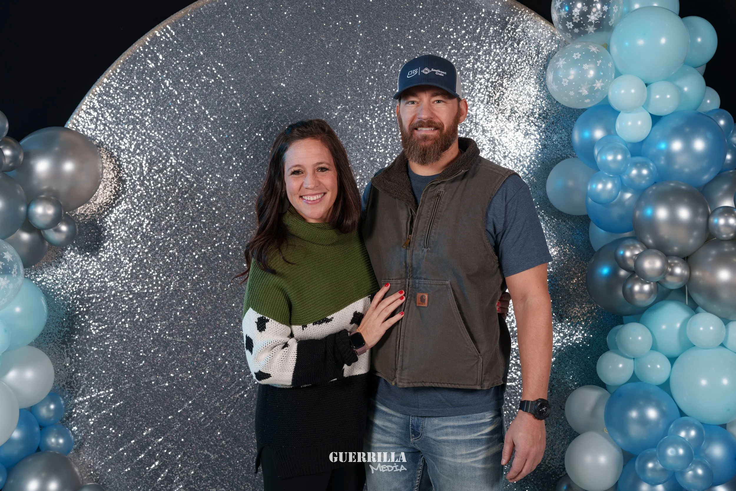 A smiling woman and man standing close together in front of a silver glitter backdrop with blue and silver balloons on each side. The woman has dark brown hair, is wearing a green, white, and black sweater, and has her hand on the man's chest. The ma