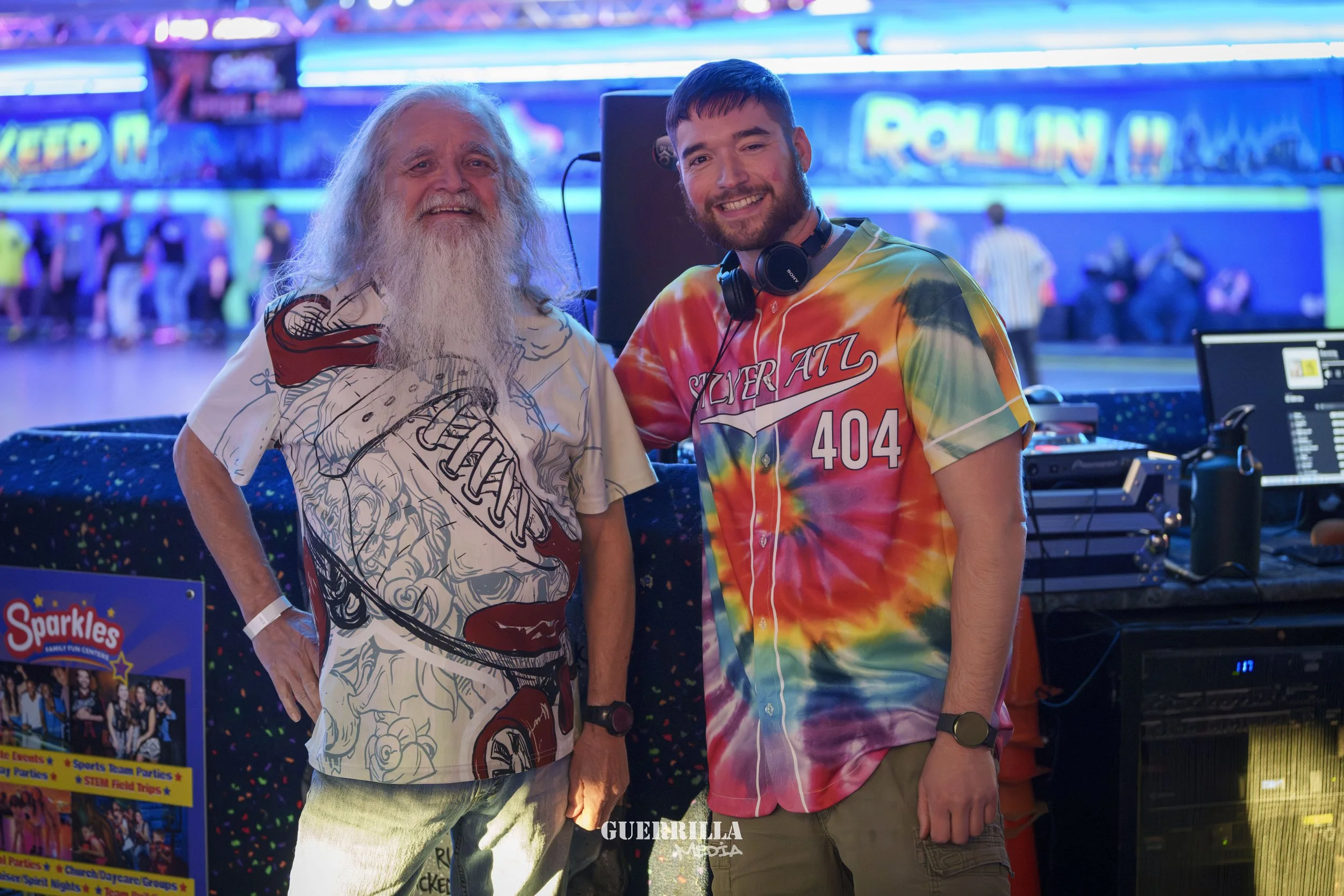 Two men smiling at a roller skating rink; one with long gray hair and beard wearing a graphic T-shirt, the other young with short dark hair and a tie-dye shirt with the number 404.