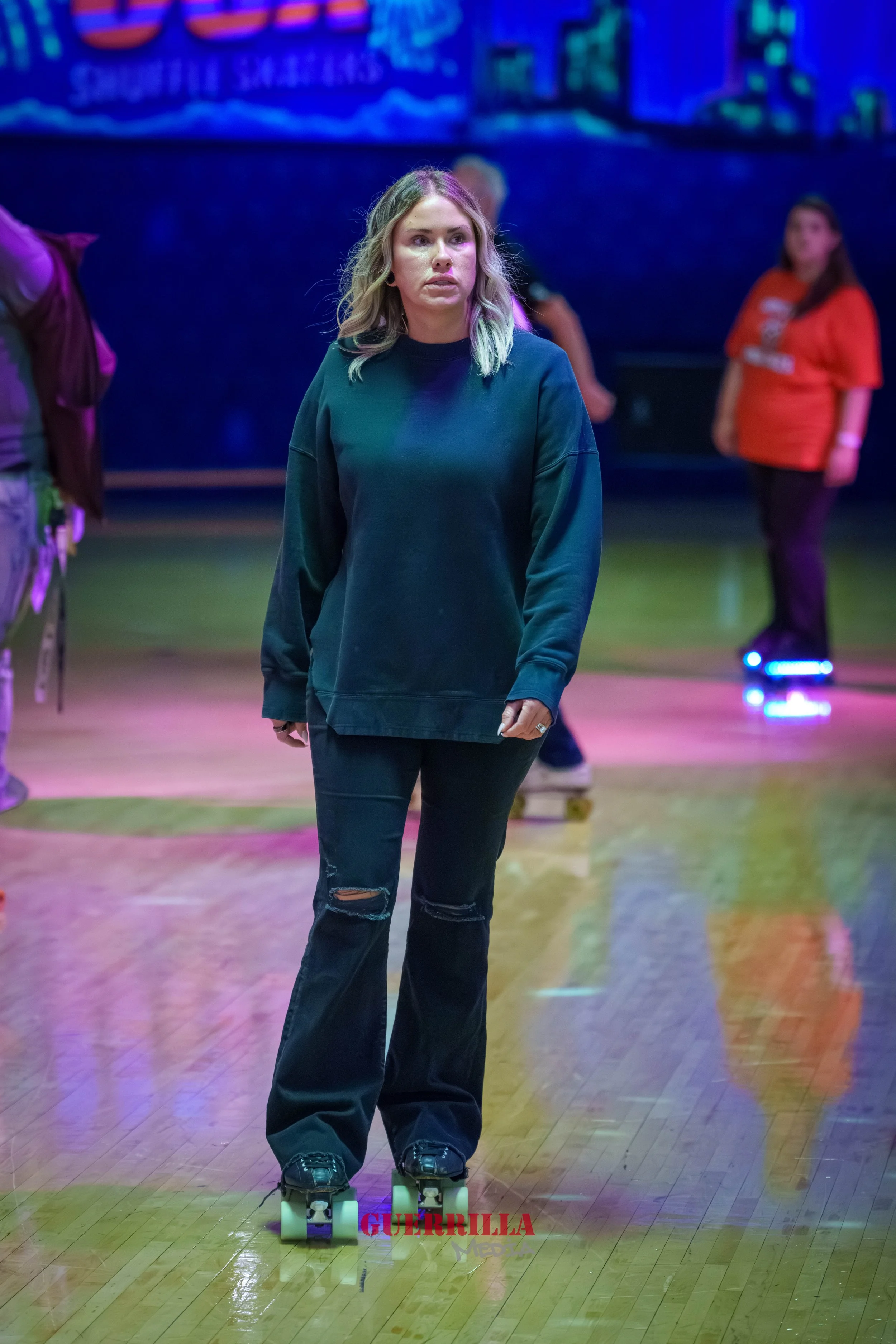 A woman with blonde hair skating on a roller rink. She wears a dark sweatshirt and ripped jeans. The background shows other people also skating and colorful neon lights.