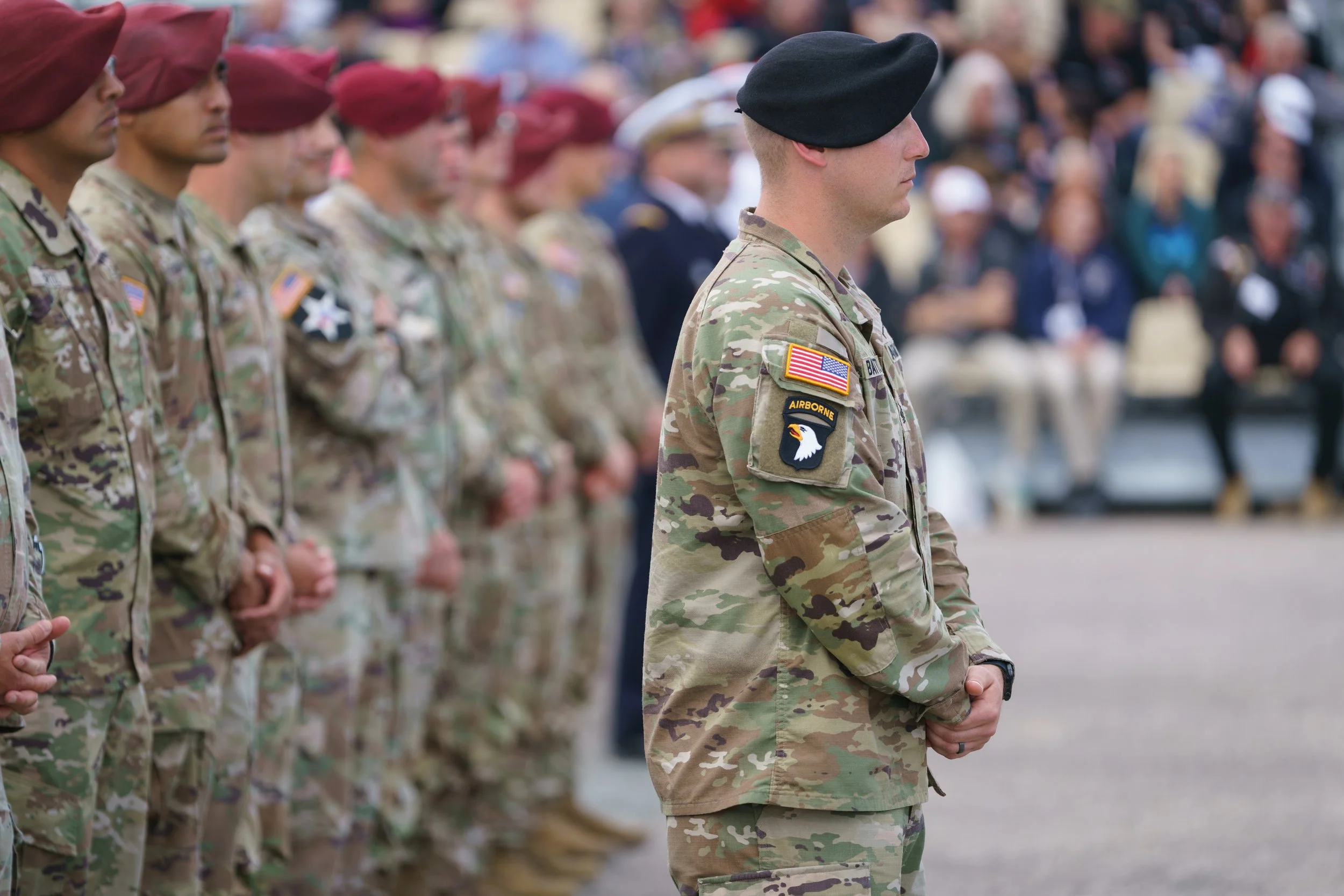 U.S. military personnel standing in formation during a ceremony, dressed in camouflage uniforms, with one soldier in the foreground wearing a black beret and a patch with an eagle emblem.