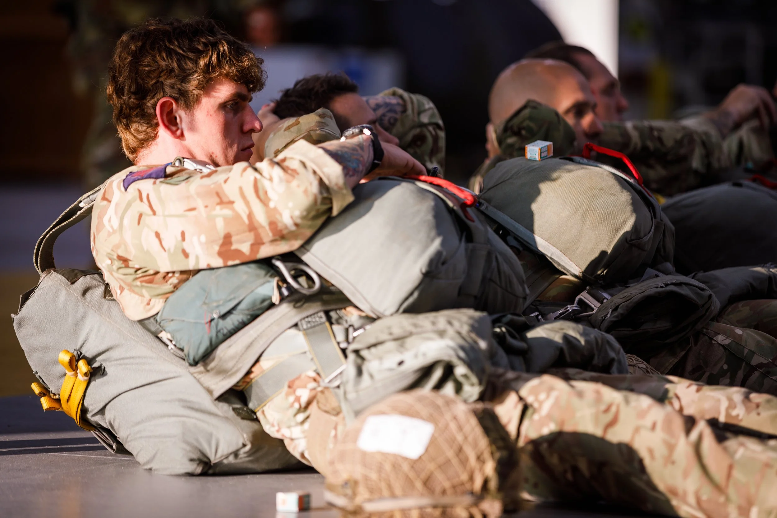 Military personnel lying on the ground with their arms crossed over their chests, dressed in camouflage uniforms and tactical gear, appearing to be in a moment of rest or reflection.