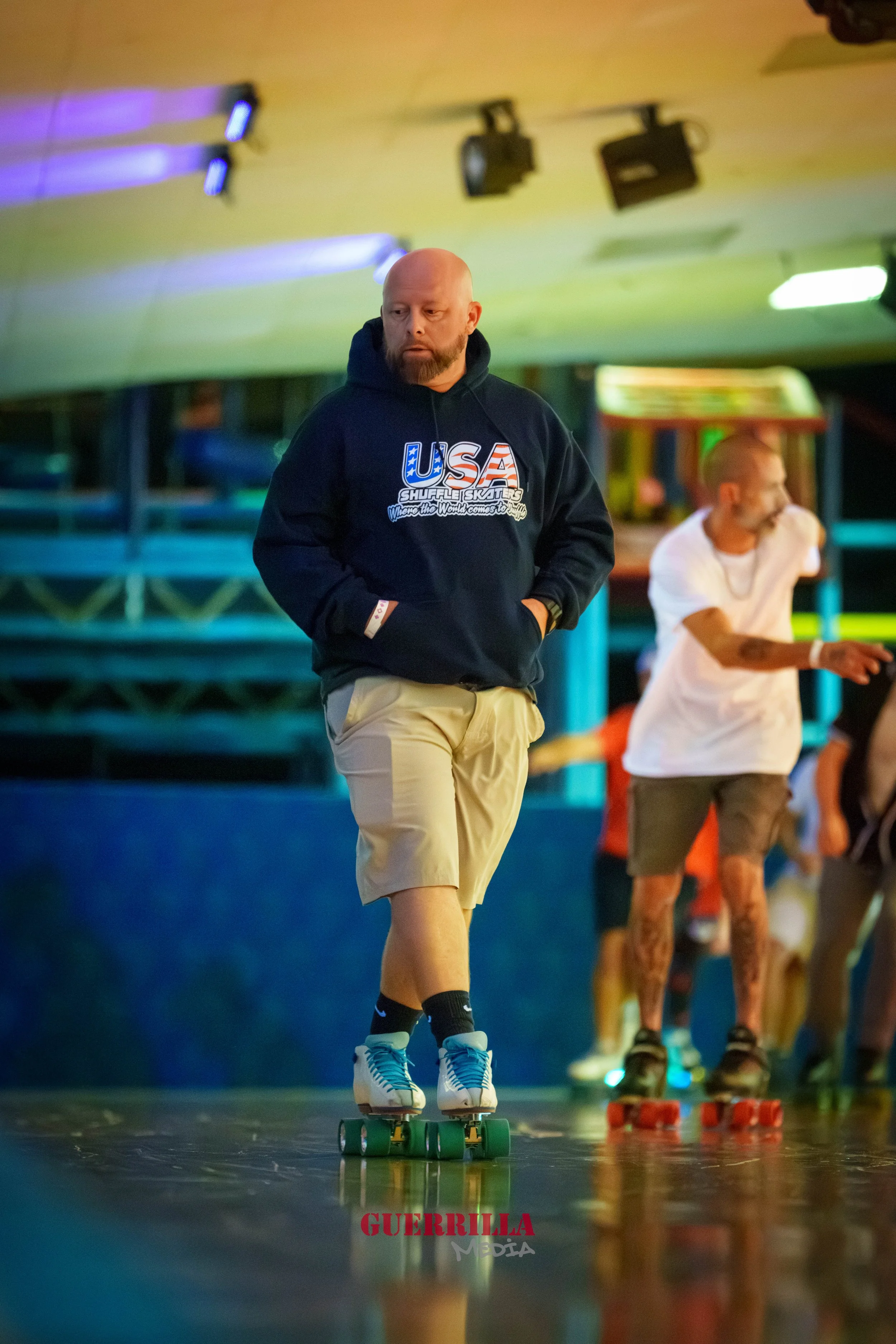Man with a beard and bald head skateboarding indoors, wearing a dark hoodie with 'USA' on it and khaki shorts, with another man skateboarding in the background.