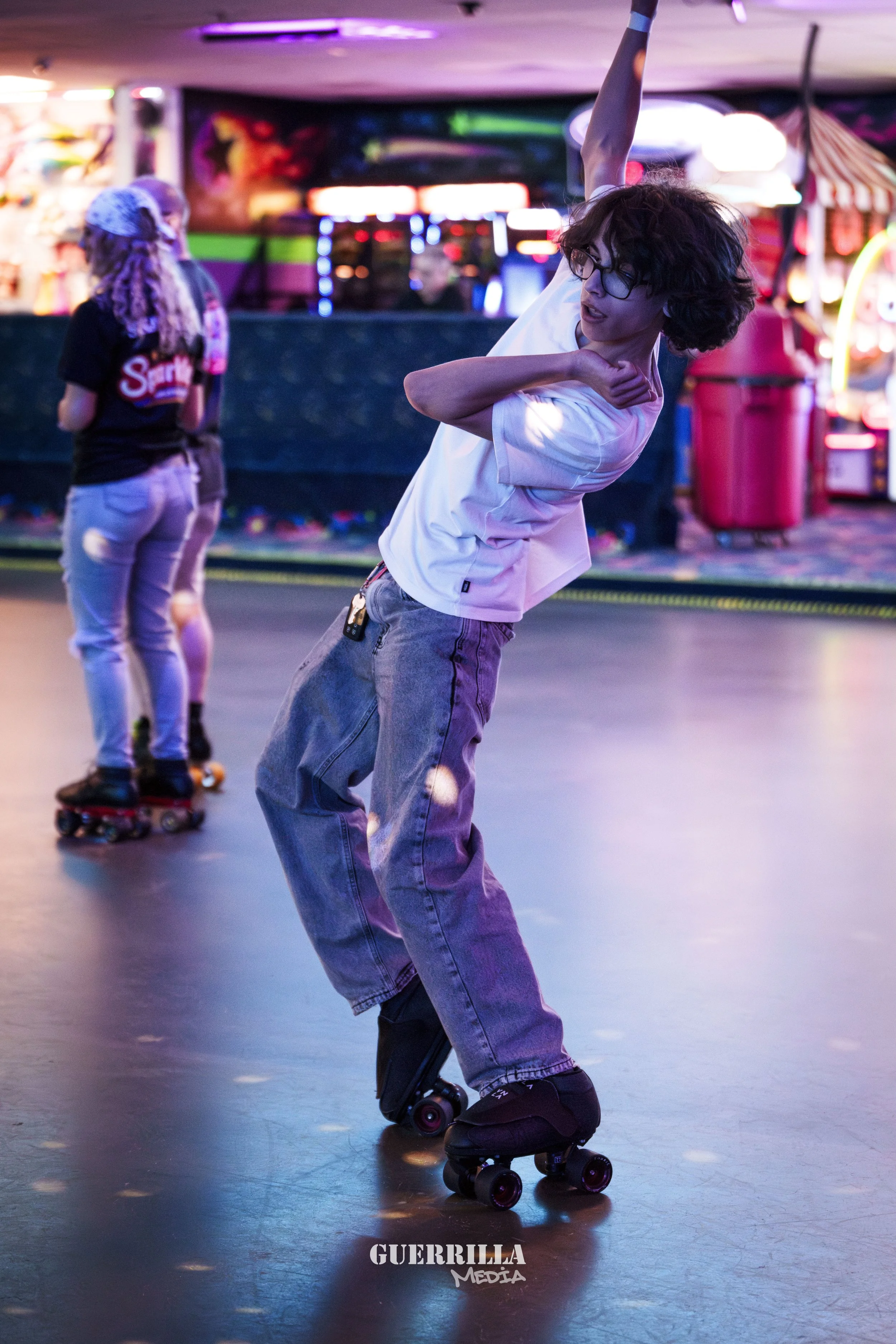 A young person with glasses skating under colorful neon lights in an arcade, with other skaters in the background.
