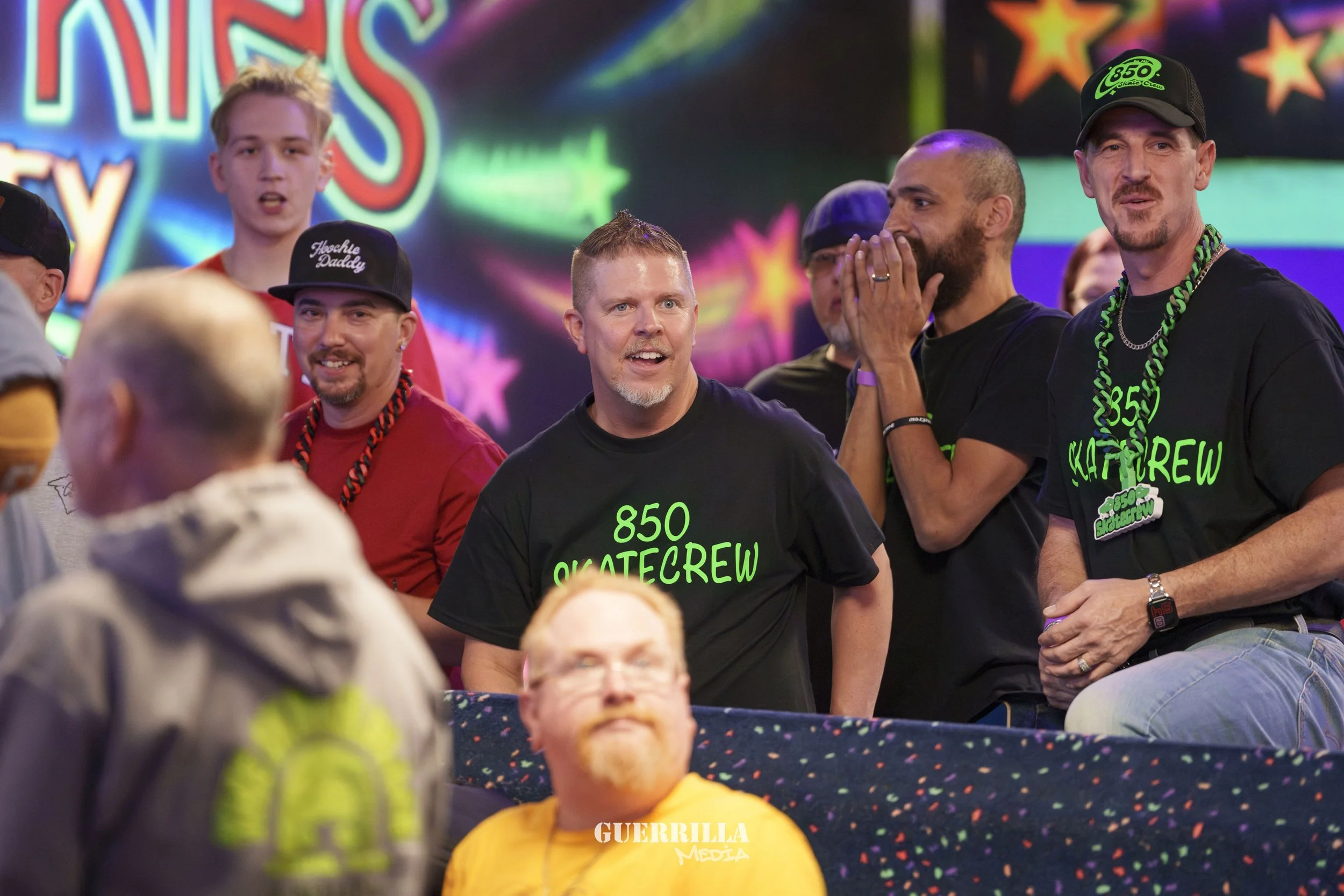 Group of people watching an event, some wearing black T-shirts with '850 SKATECREW' printed in green, colorful background with stars, and a mix of expressions.