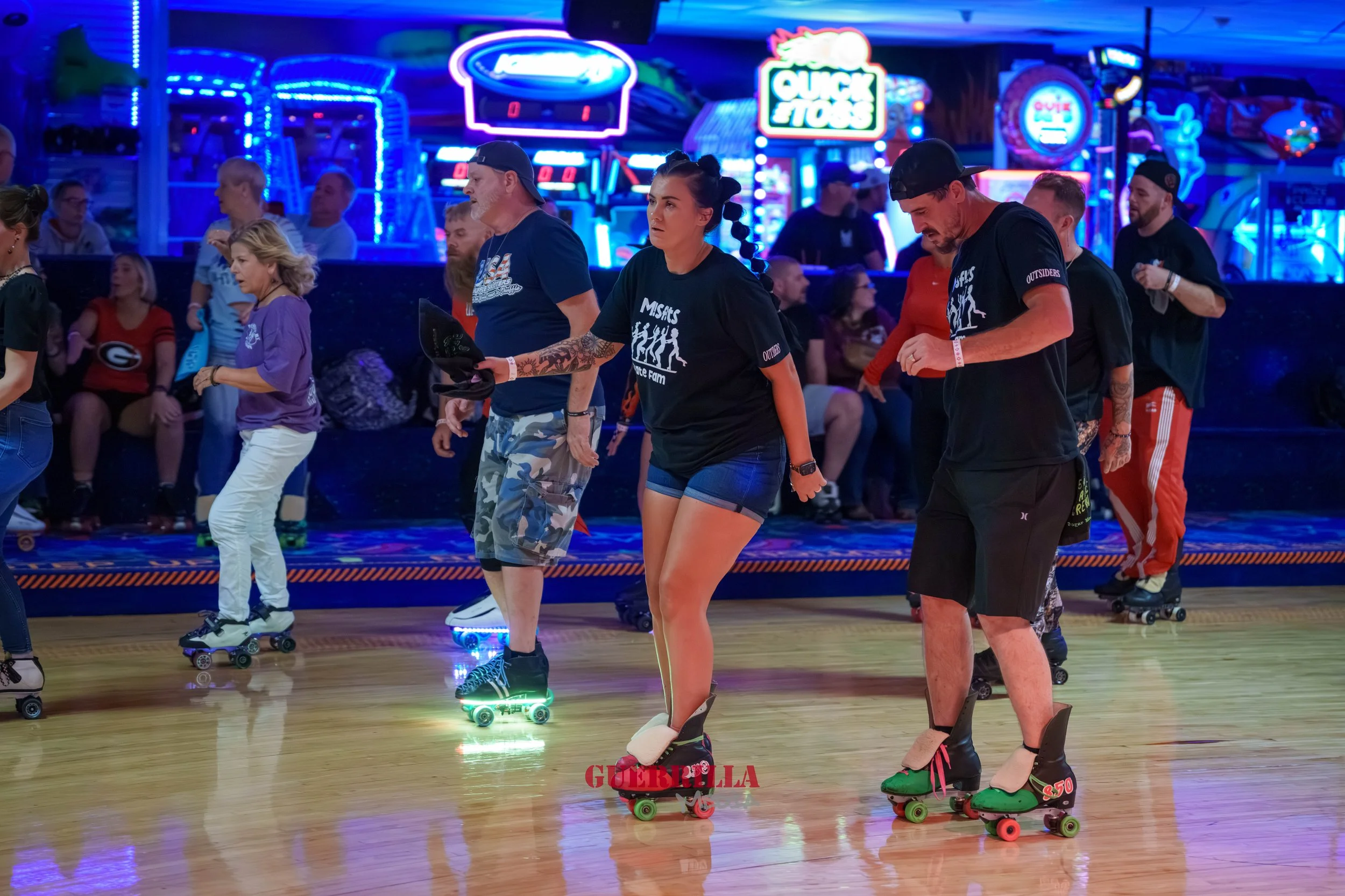 People roller skating in an indoor roller rink with neon lights and signs, including a "Quick Toss" arcade game, and some spectators sitting in the background.