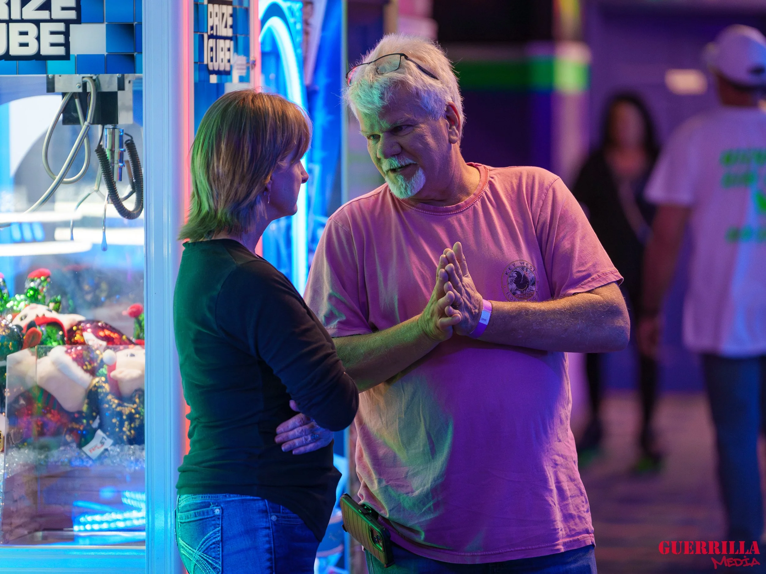Two people are having a conversation at a carnival game booth, with neon lights and stuffed animals in the background, while other people walk in the background.