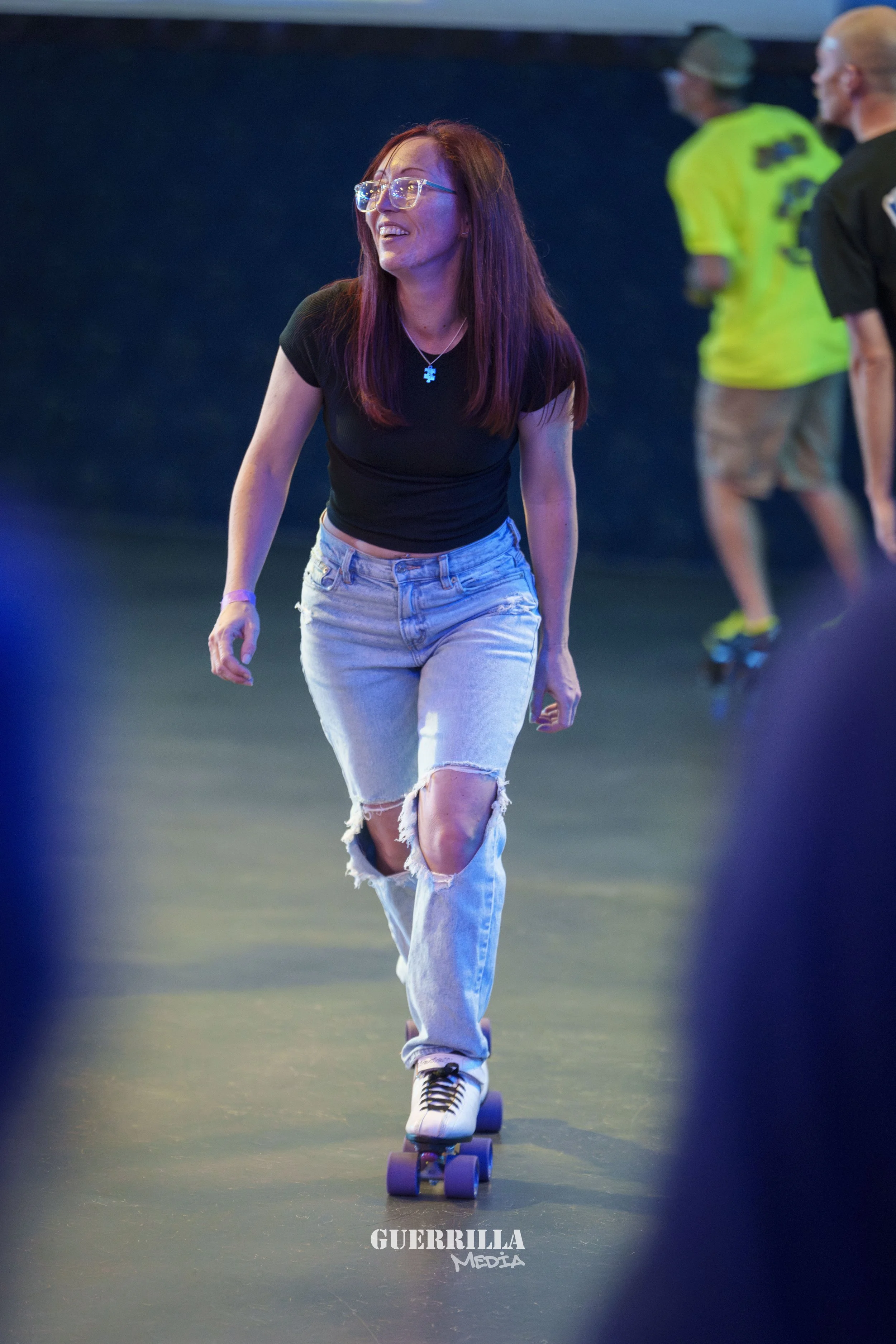 A young woman with red hair and glasses is skateboarding indoors, wearing a black t-shirt and ripped jeans. She is smiling and appears to be enjoying herself.