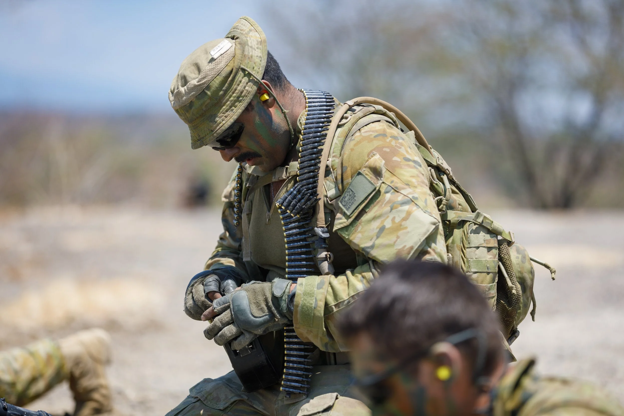 A soldier in camouflage uniform, wearing a helmet and protective gear, wearing ammo belts, and preparing equipment outdoors during the day.
