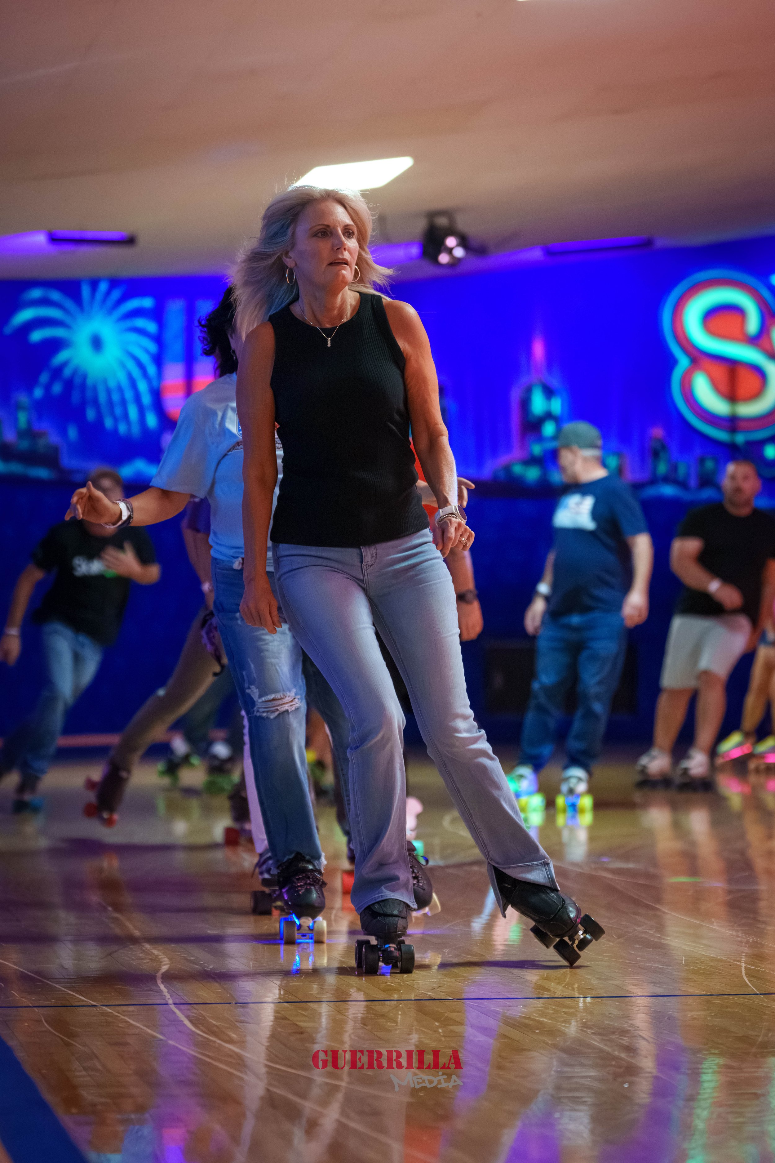 People roller-skating on an indoor rink with colorful neon lights and cityscape artwork on the walls.
