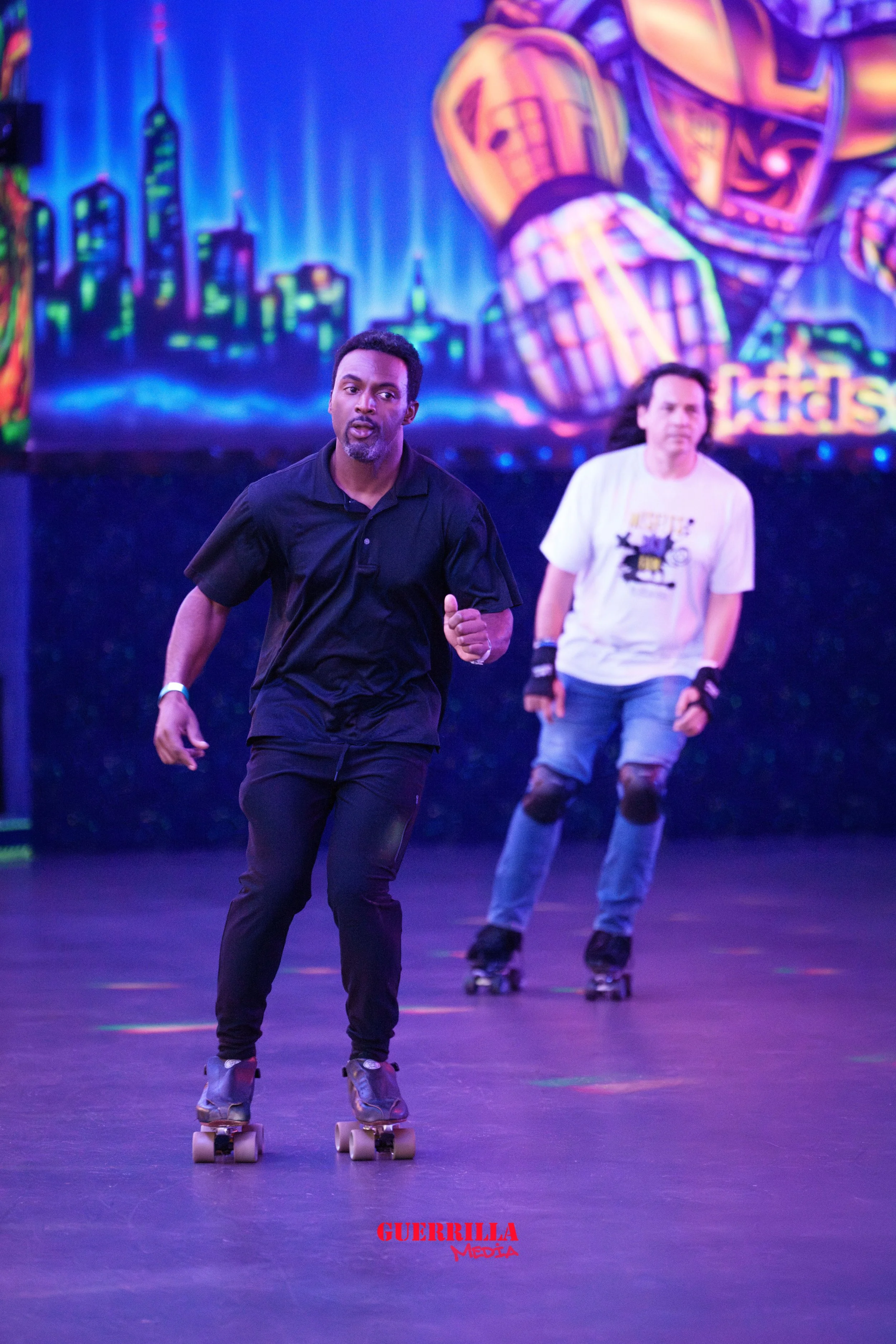 Two men roller skating indoors, with a colorful cityscape mural in the background. The man in front is wearing a black shirt and black pants, and the man behind is wearing a white t-shirt and distressed jeans with knee pads.