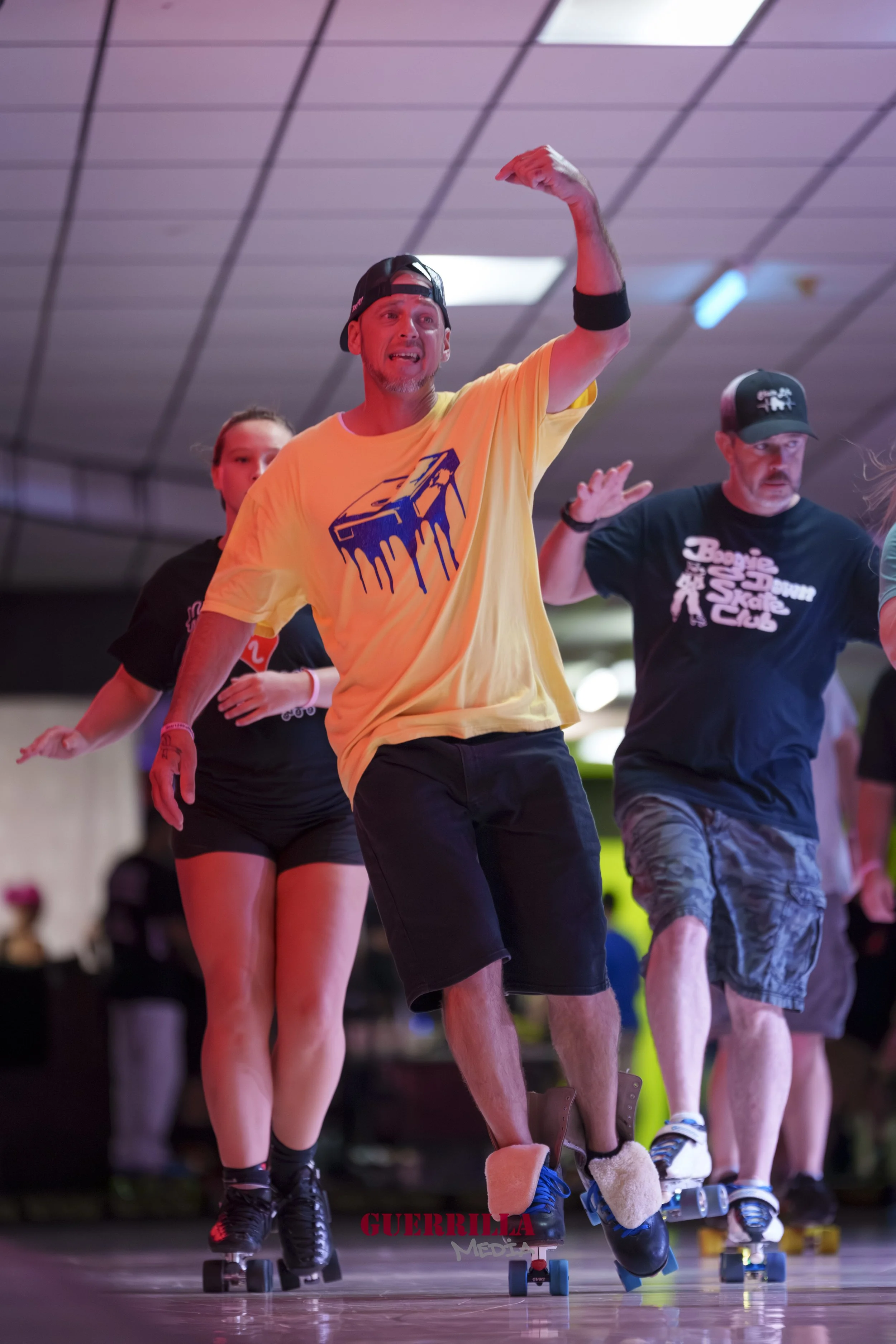 People roller skating indoors, with a man in a yellow shirt, black shorts, and a black cap leading others. The man appears to be raising his right arm and has a cloth or band on his left arm. There is a woman and another man, also roller skating, in 