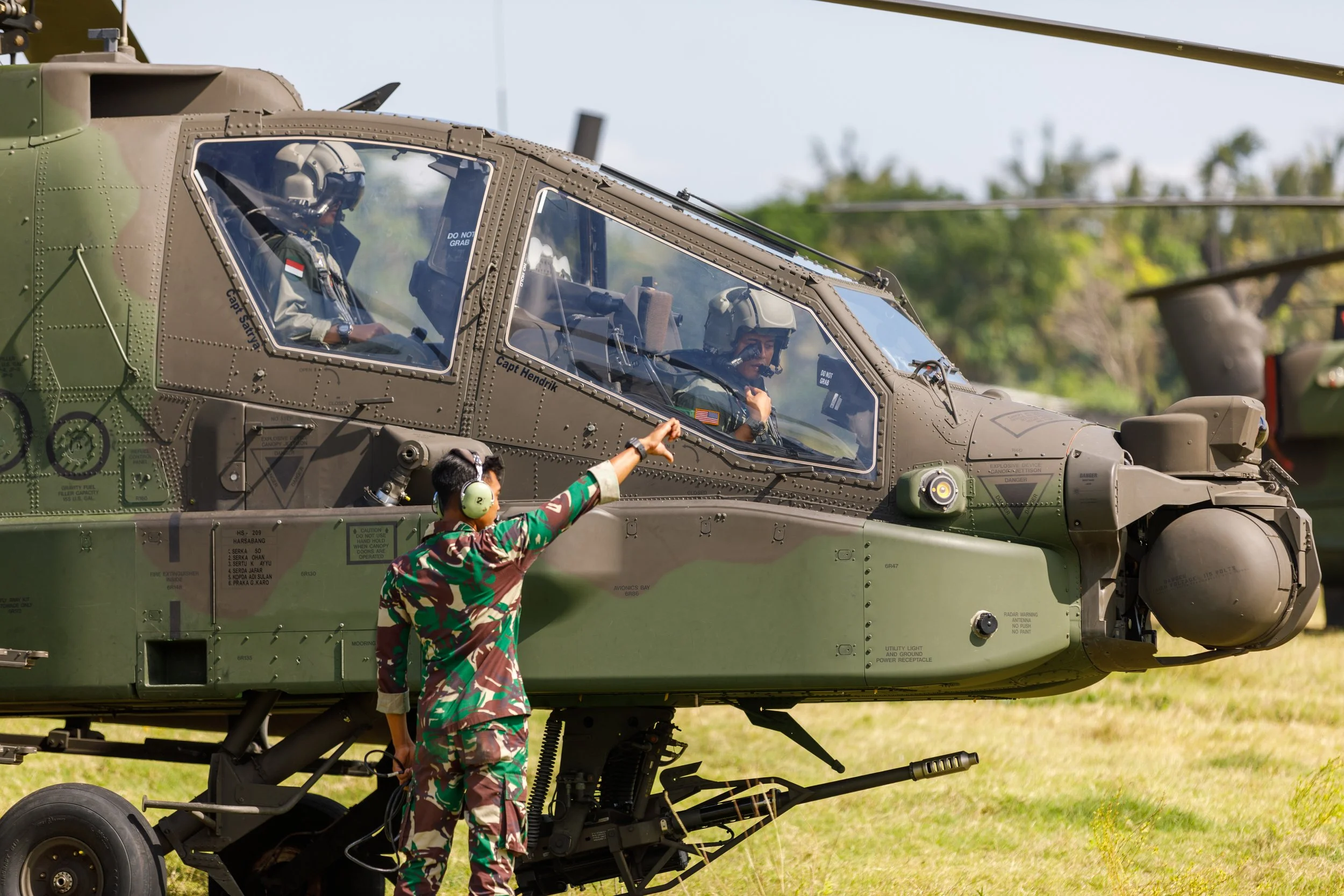 A soldier in camouflage uniform wearing headphones is pointing at the cockpit of a military helicopter on a grassy field, with two pilots inside the helicopter.