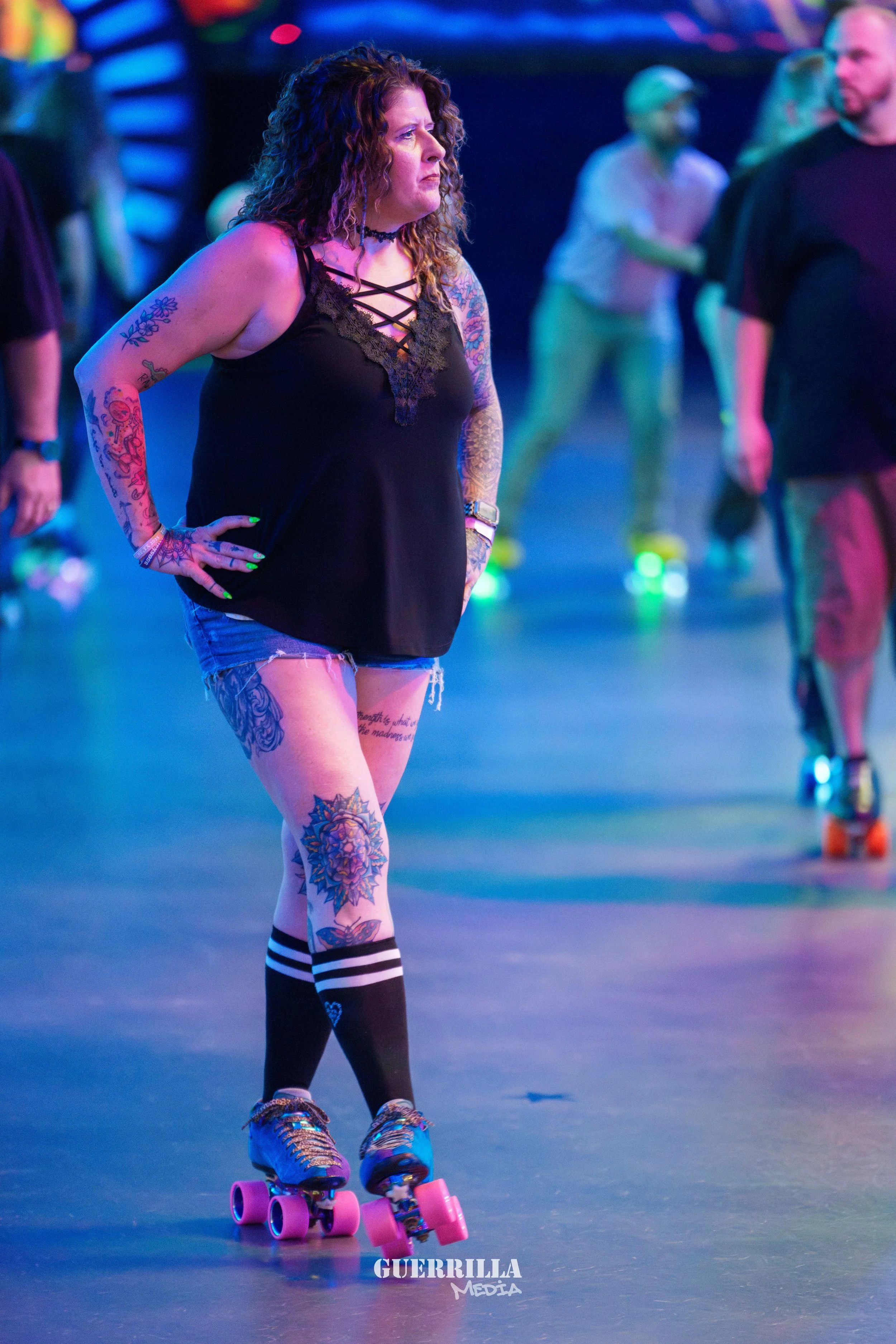 A woman with curly hair and tattoos roller skating indoors with other people in the background under colorful lighting.
