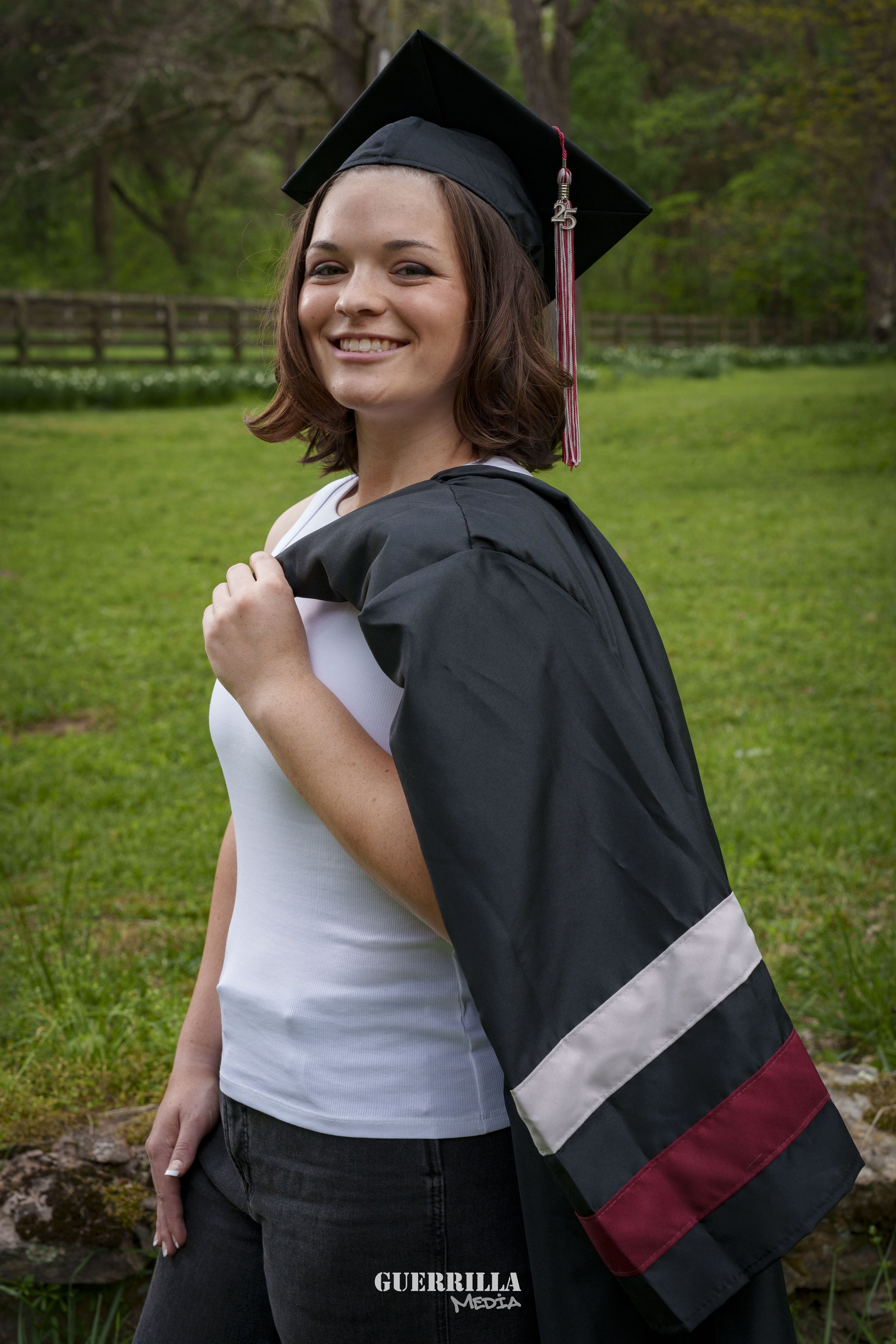 A young woman smiling outdoors in a grassy area, wearing a black graduation cap and gown draped over her shoulder, with trees and a wooden fence in the background.
