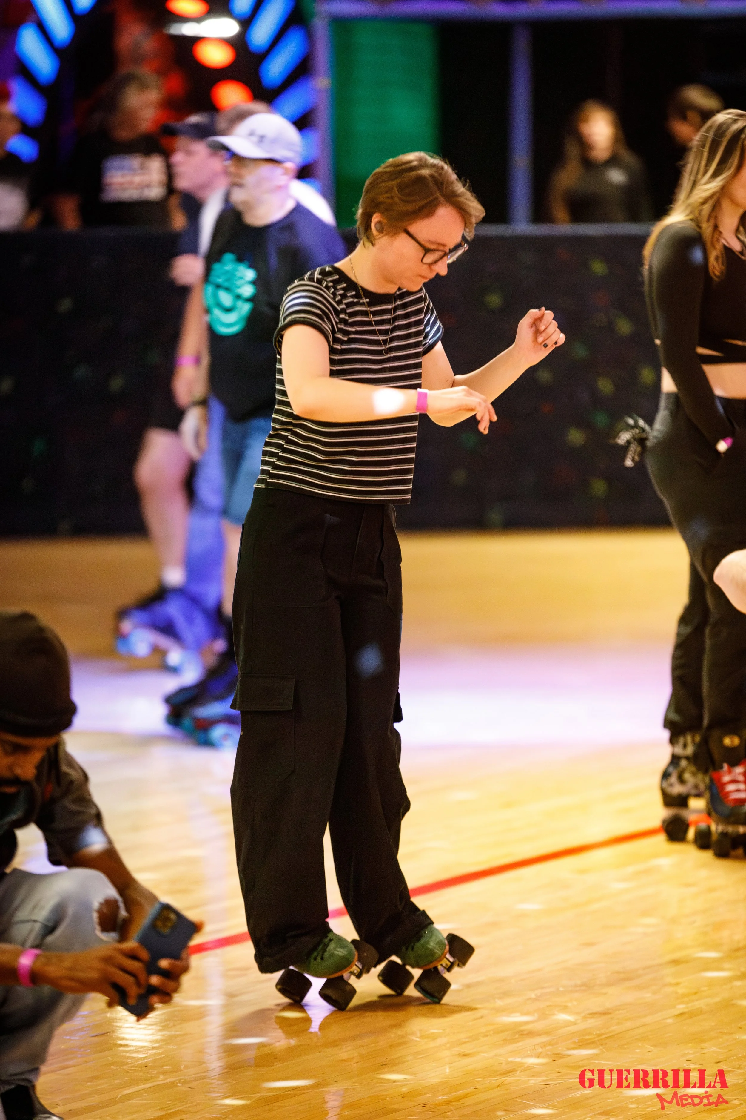 A young woman roller skating indoors on a wooden floor, wearing glasses, a black and white striped t-shirt, and black cargo pants. She is looking down and adjusting her roller skate. There are other people in the background, some wearing casual cloth