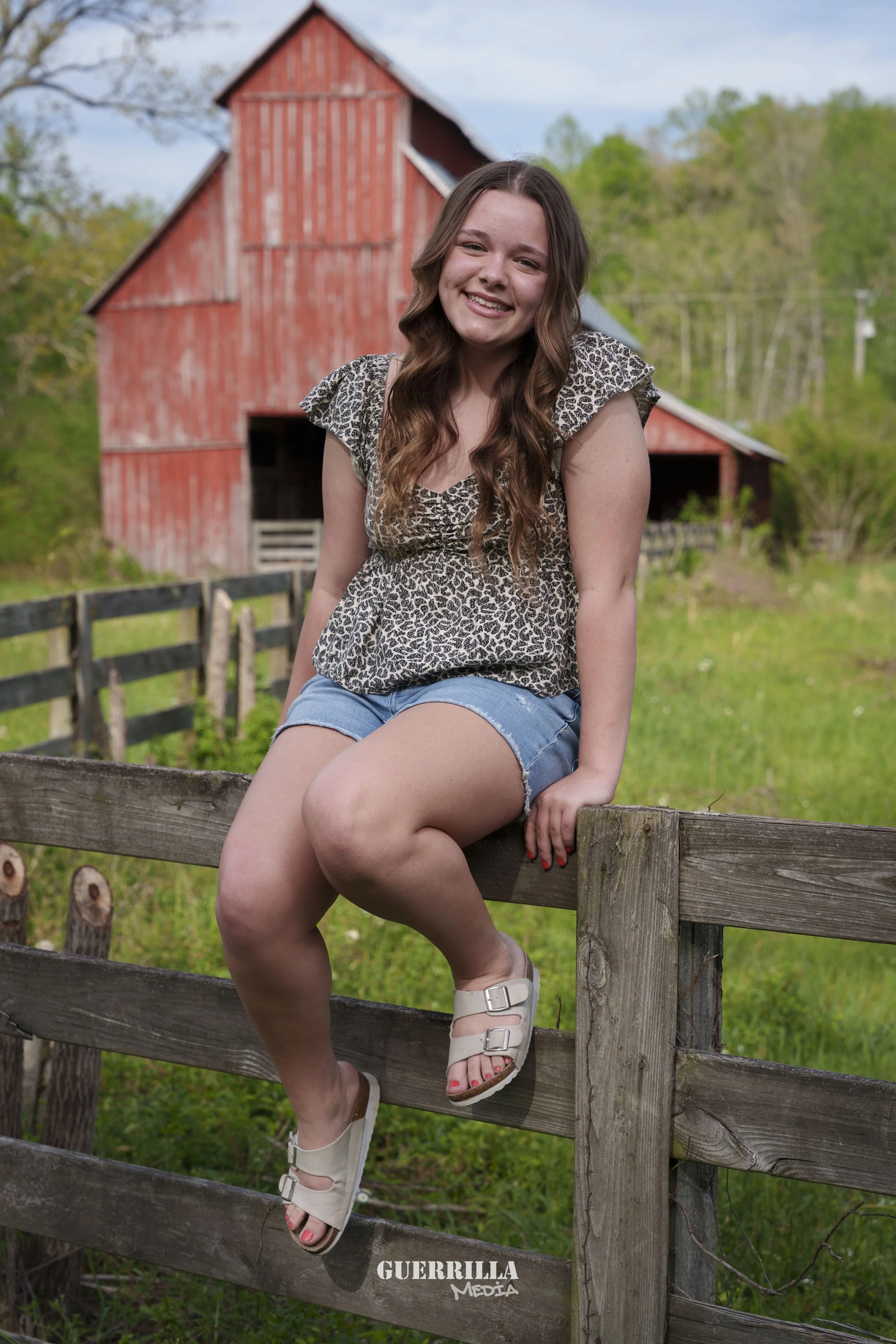 A young woman with long brown wavy hair smiling and sitting on a wooden fence in front of a red barn, wearing a leopard print top and denim shorts.