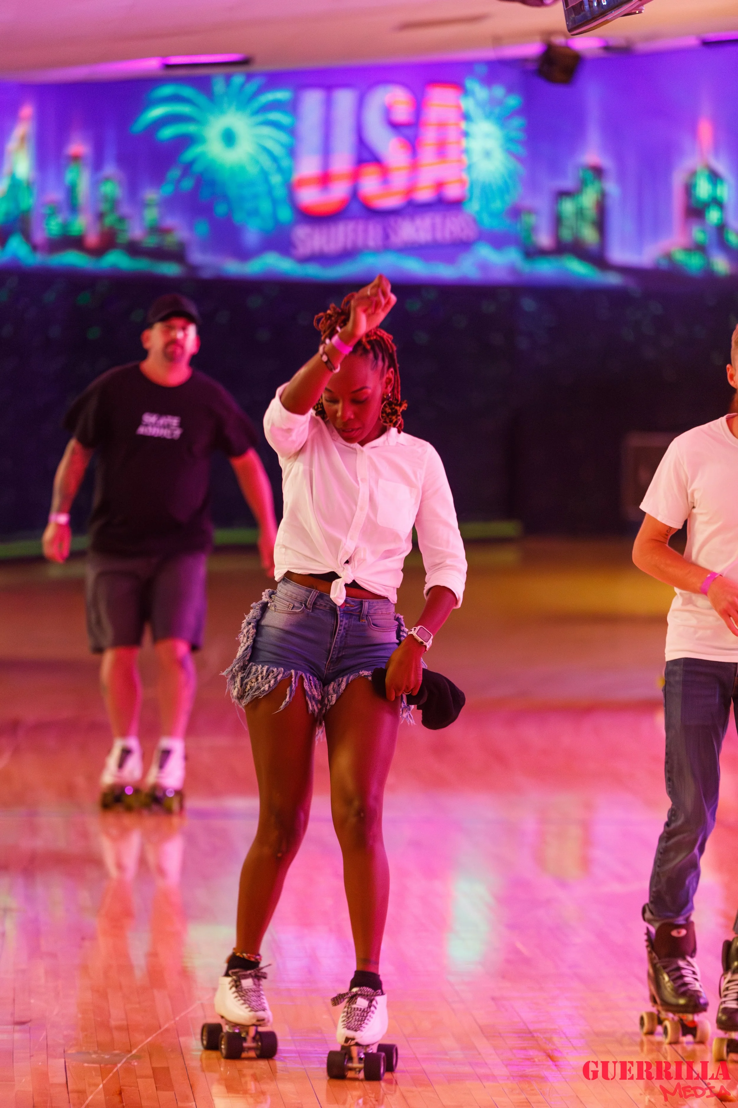 A woman roller skating on a wooden floor at an indoor skating rink with vibrant neon lighting and a large screen in the background displaying the word 'USA' and fireworks.