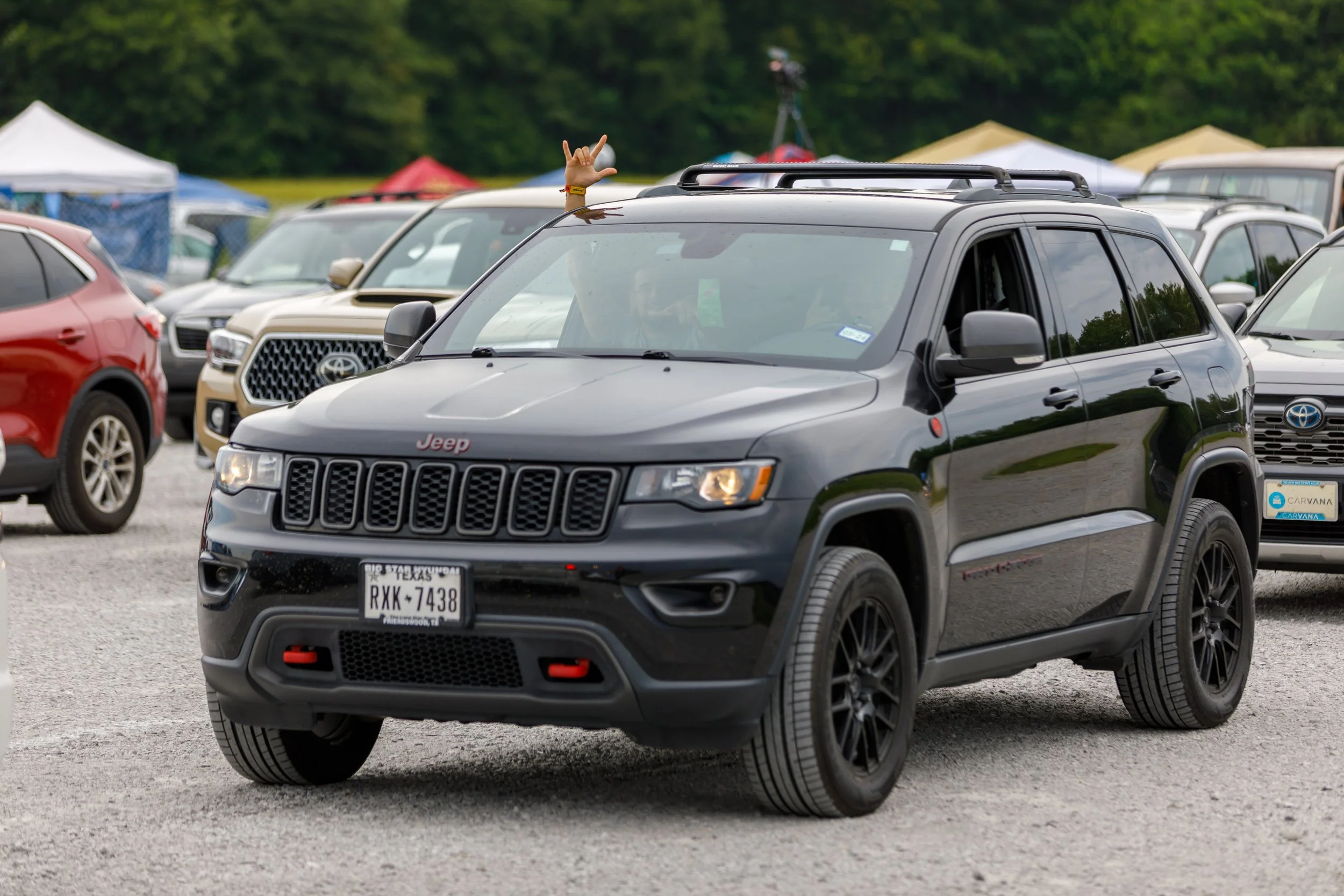 Black Jeep SUV with a person inside making a hand gesture, parked among other vehicles in a lot with tents in the background.