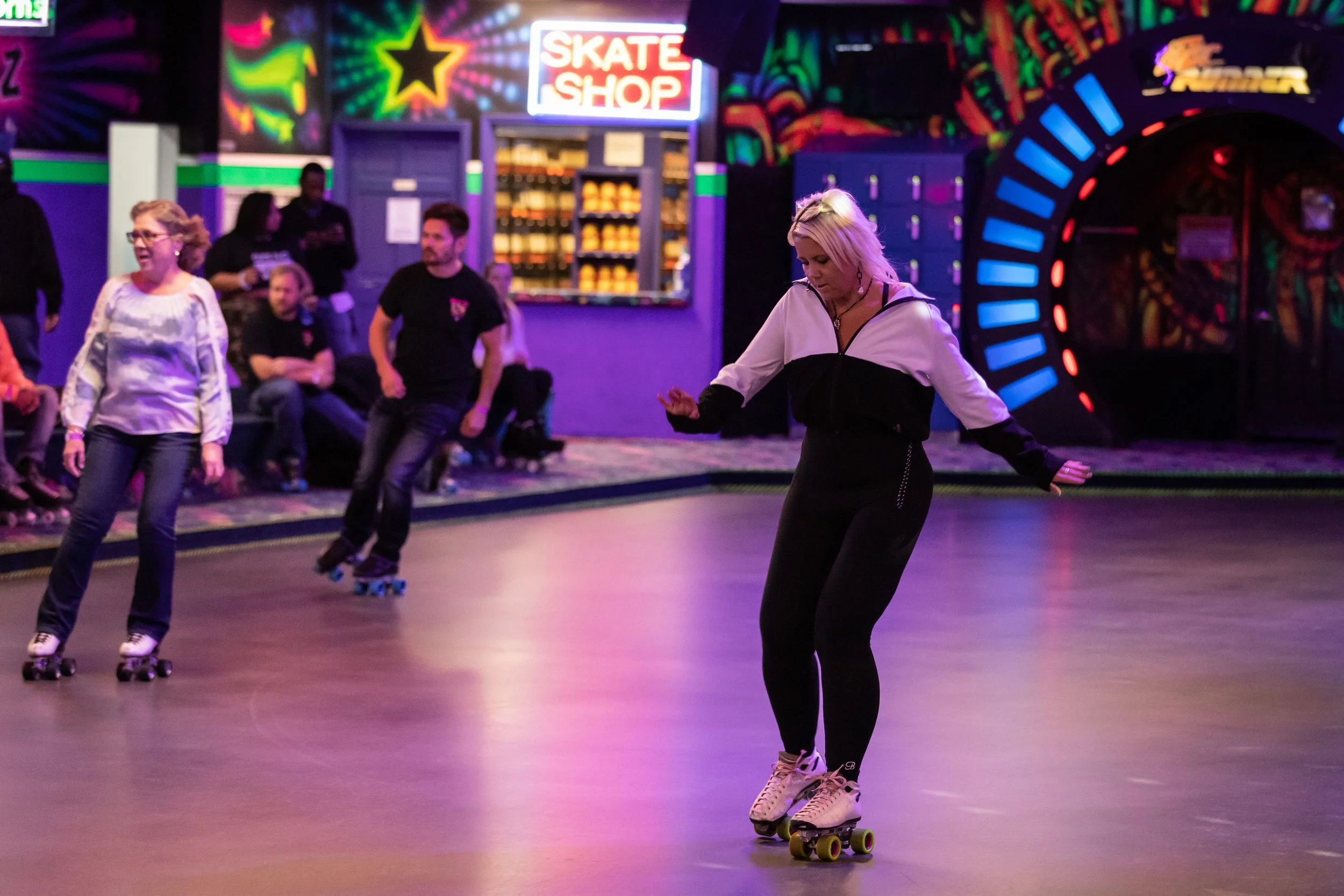 Five people roller skating in a colorful indoor roller rink with neon lights, a skate shop, and arcade games in the background.
