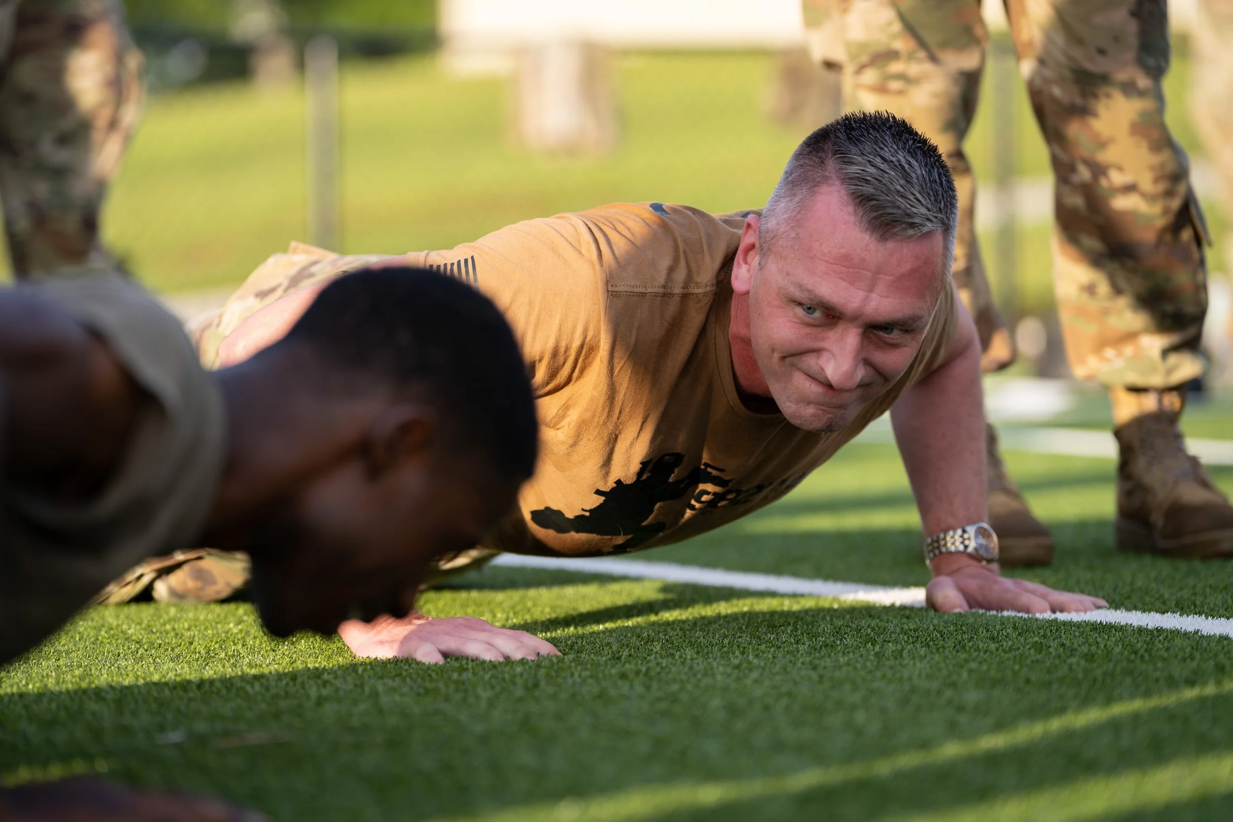 A man in military uniform doing push-ups outdoors while others in uniform stand around him.