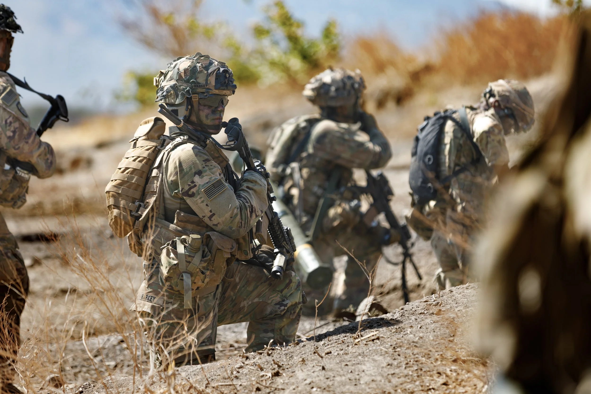Military soldiers in camouflage gear kneeling and standing with weapons in a desert environment.