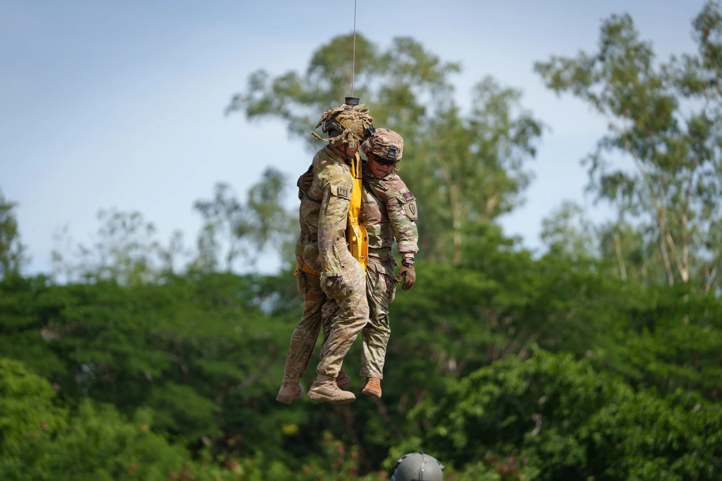Two soldiers in camouflage uniforms are holding onto each other while jumping from a helicopter during a training exercise outdoors with trees in the background.