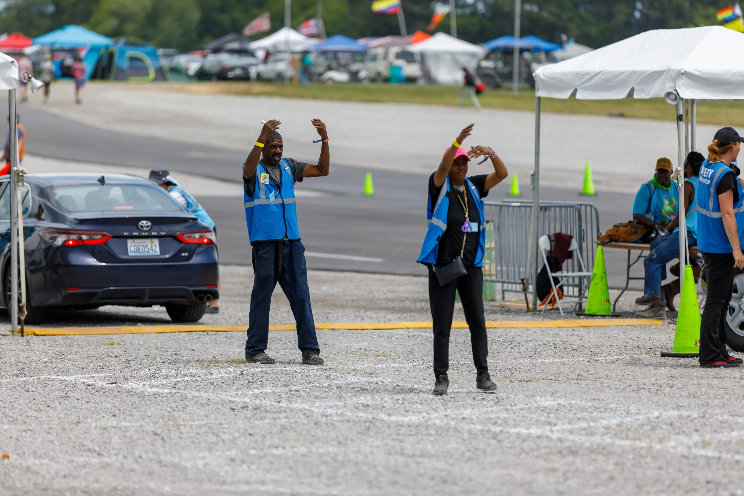 Two volunteers standing in a parking area at an outdoor event, with tents and cars in the background, waving and guiding attendees.