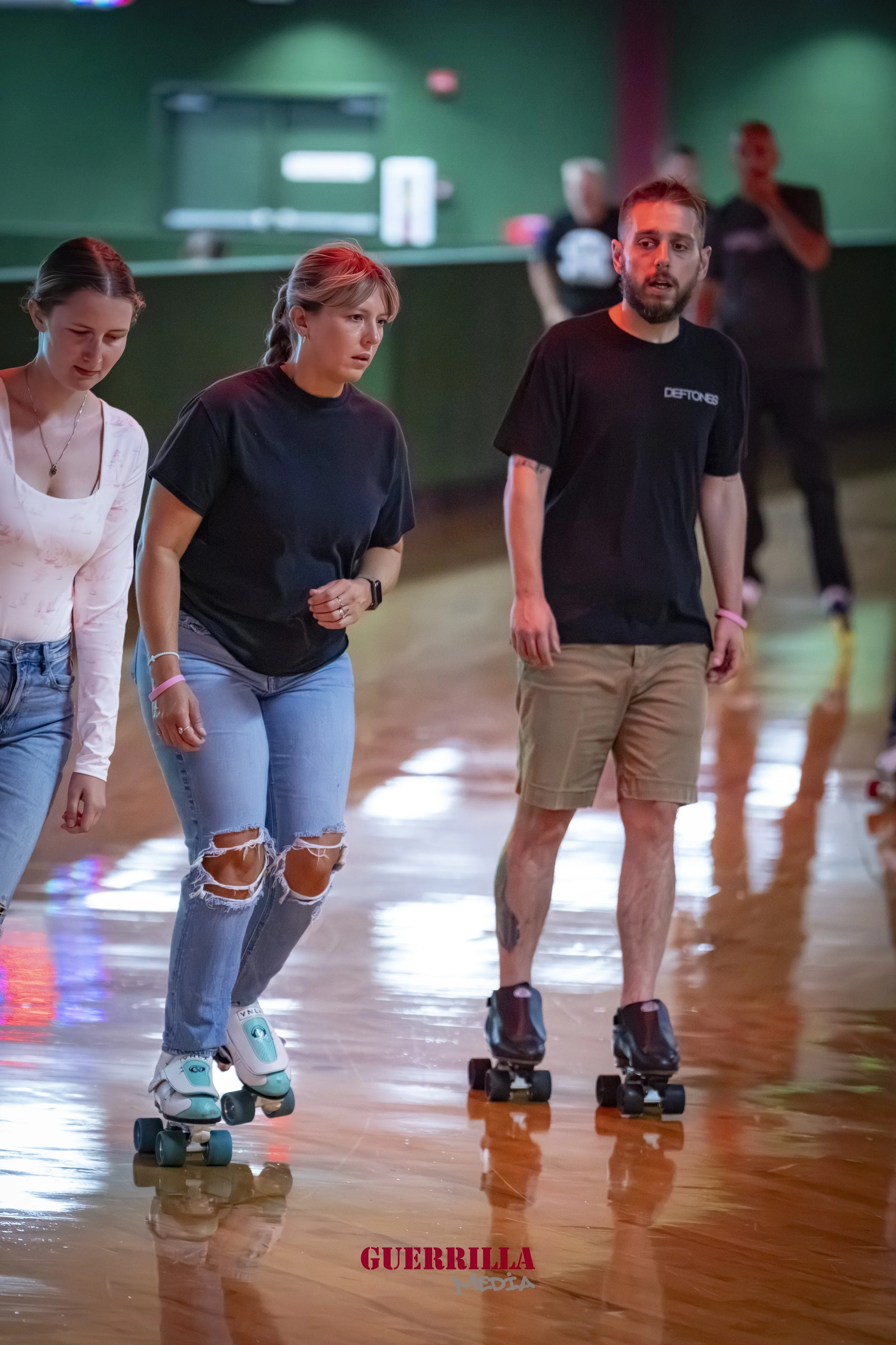 Three people roller skating indoors, two women and one man, with focused and relaxed expressions, in a facility with a polished wooden floor and green walls, and others skating in the background.