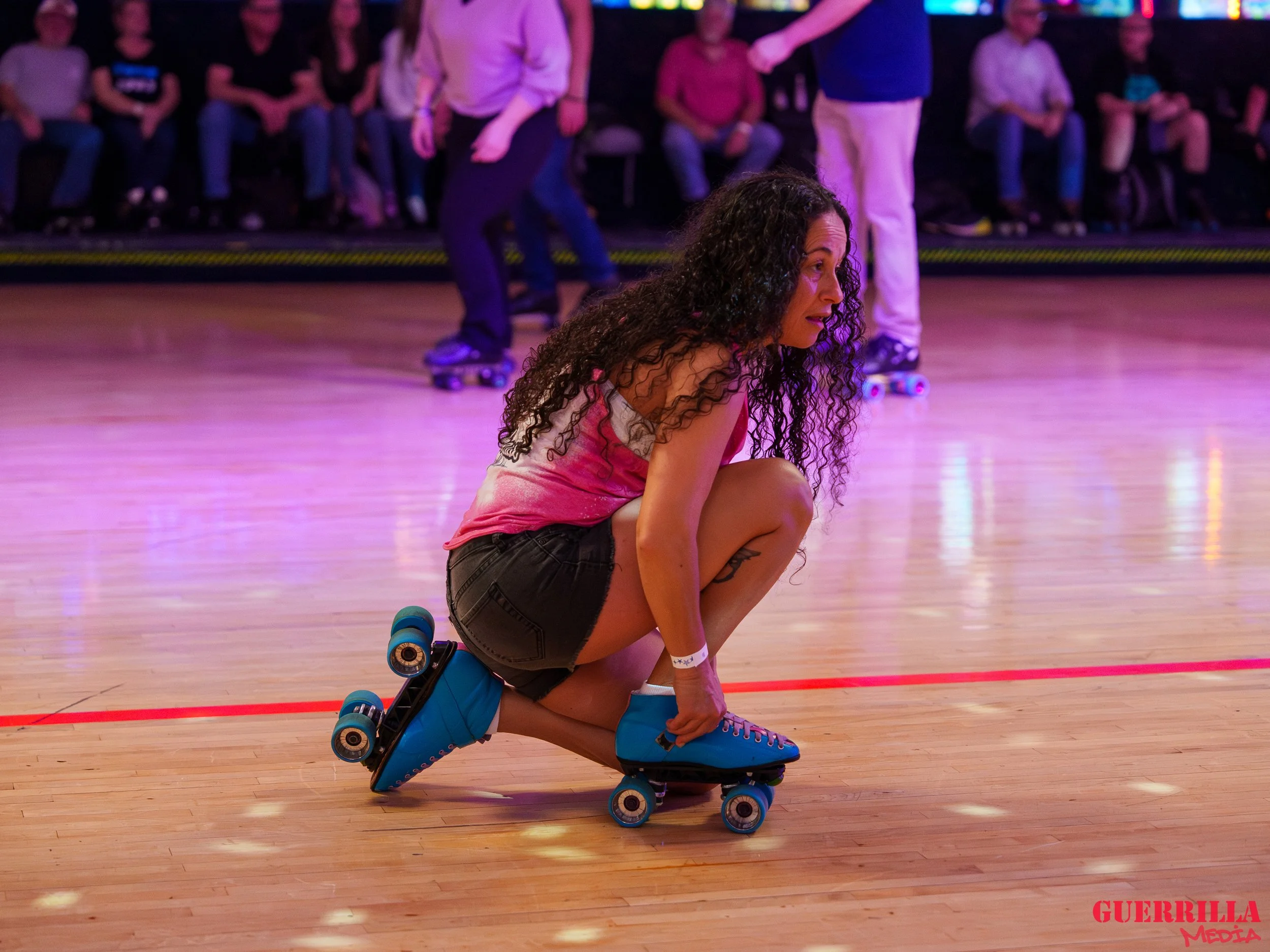 A woman in a pink tank top and black shorts crouches on a roller skate rink, holding her skate with one hand while surrounded by other skaters and spectators in the background.